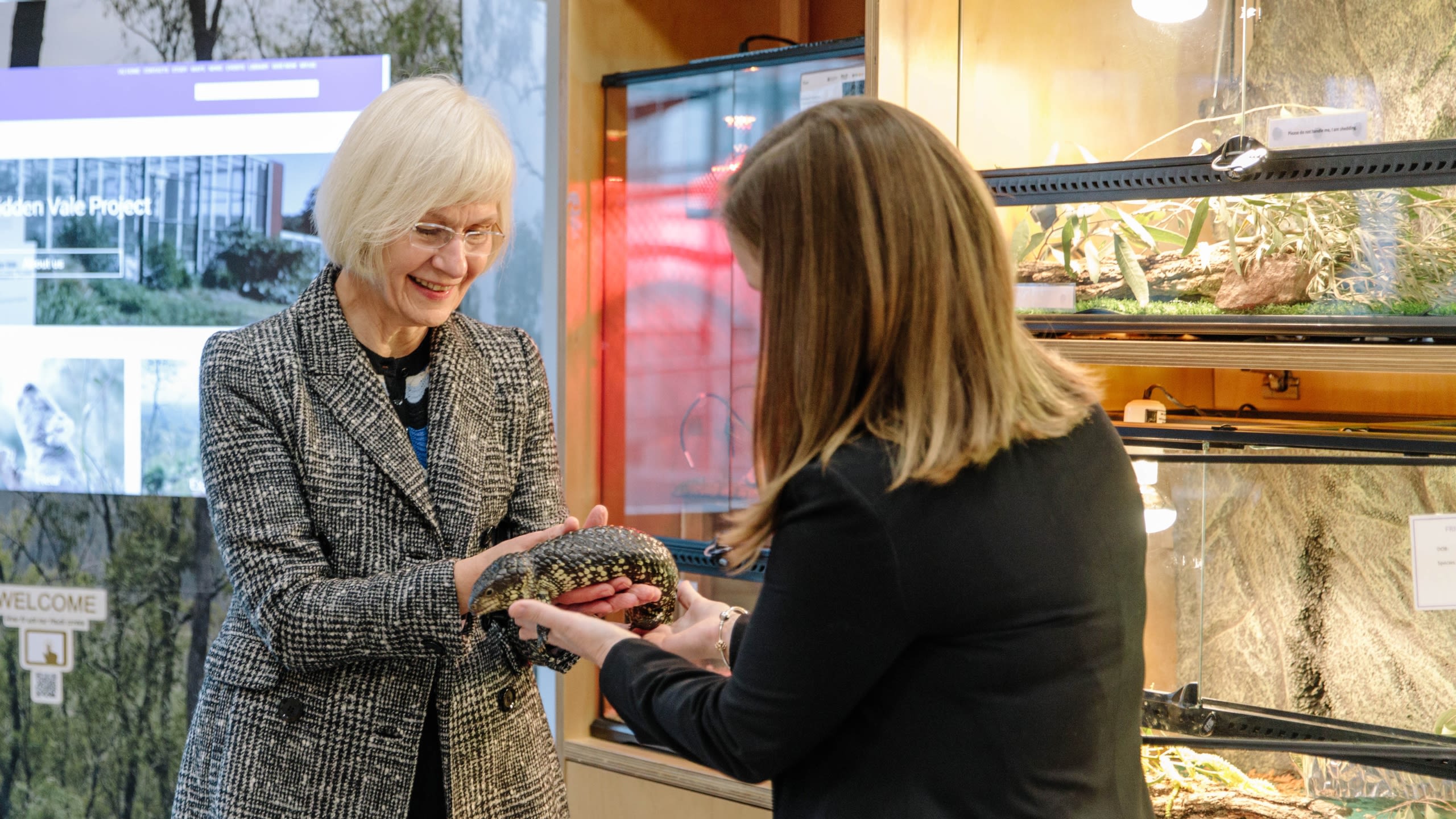 Two women hold a bobtail lizard in front of glass enclosures filled with leaves