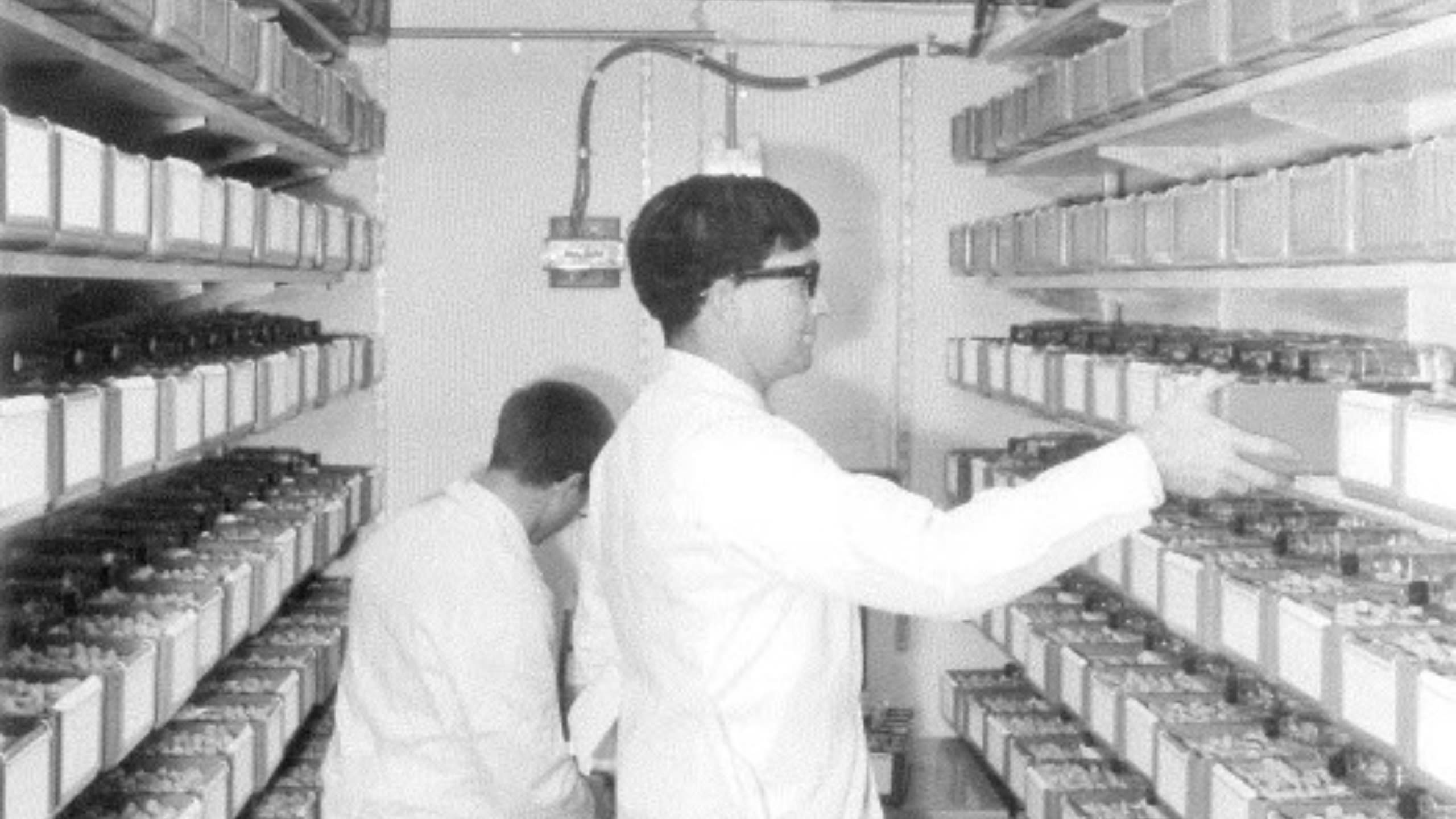 two men in white lab coats and safety glasses standing in a small room between two walls stacked floor to ceiling full of specimen containers