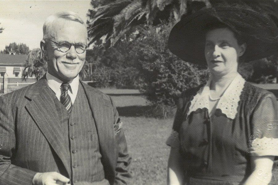A sepia image of a man in a suit and a woman in a dress with a lace collar and a large wide-brimmed hat smiling for the camera