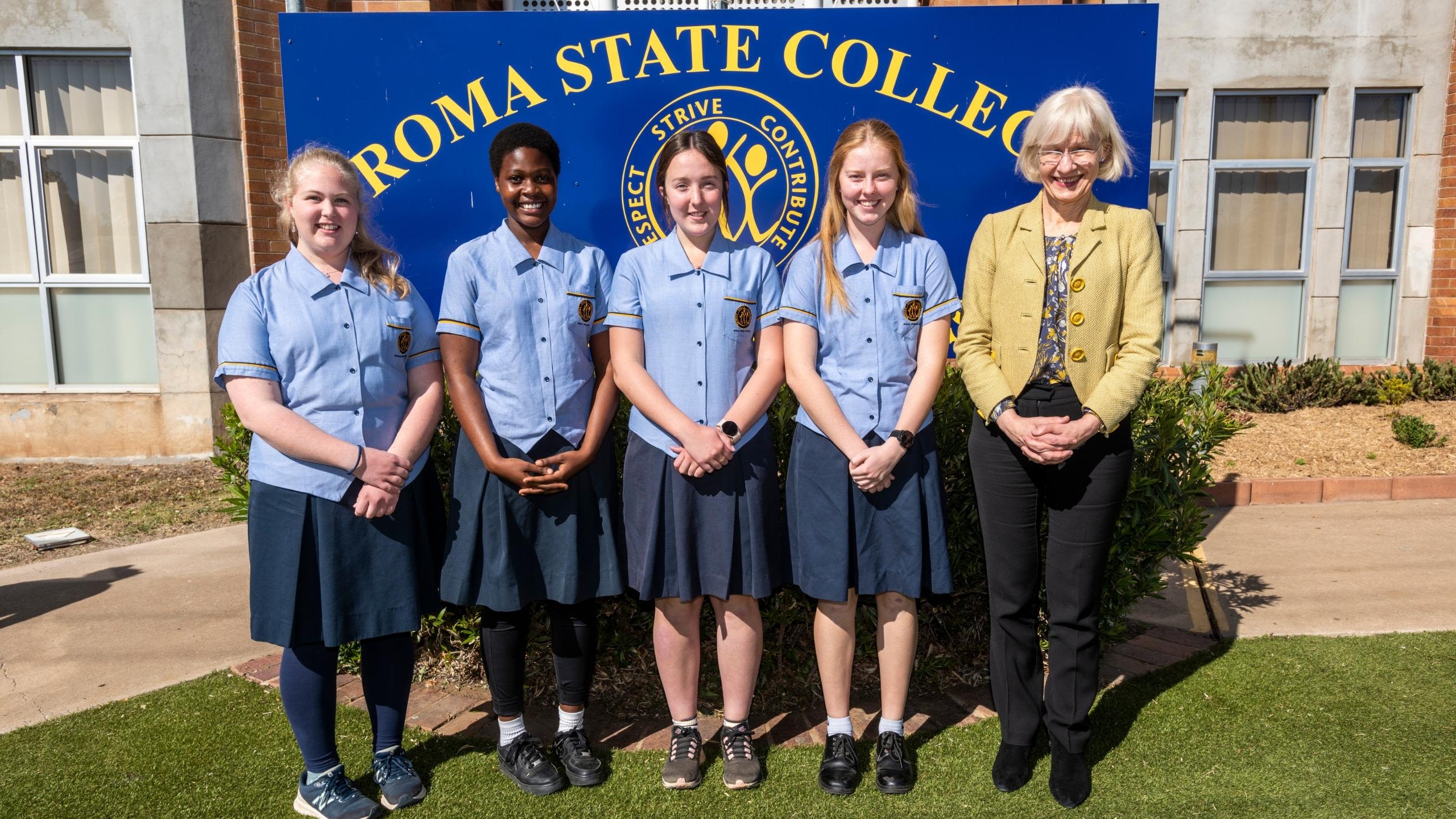 4 high school students in blue school uniforms are standing with the Vice-Chancellor from the University, who is wearing a yellow jacket. They are standing in front of the large Roma State College sign