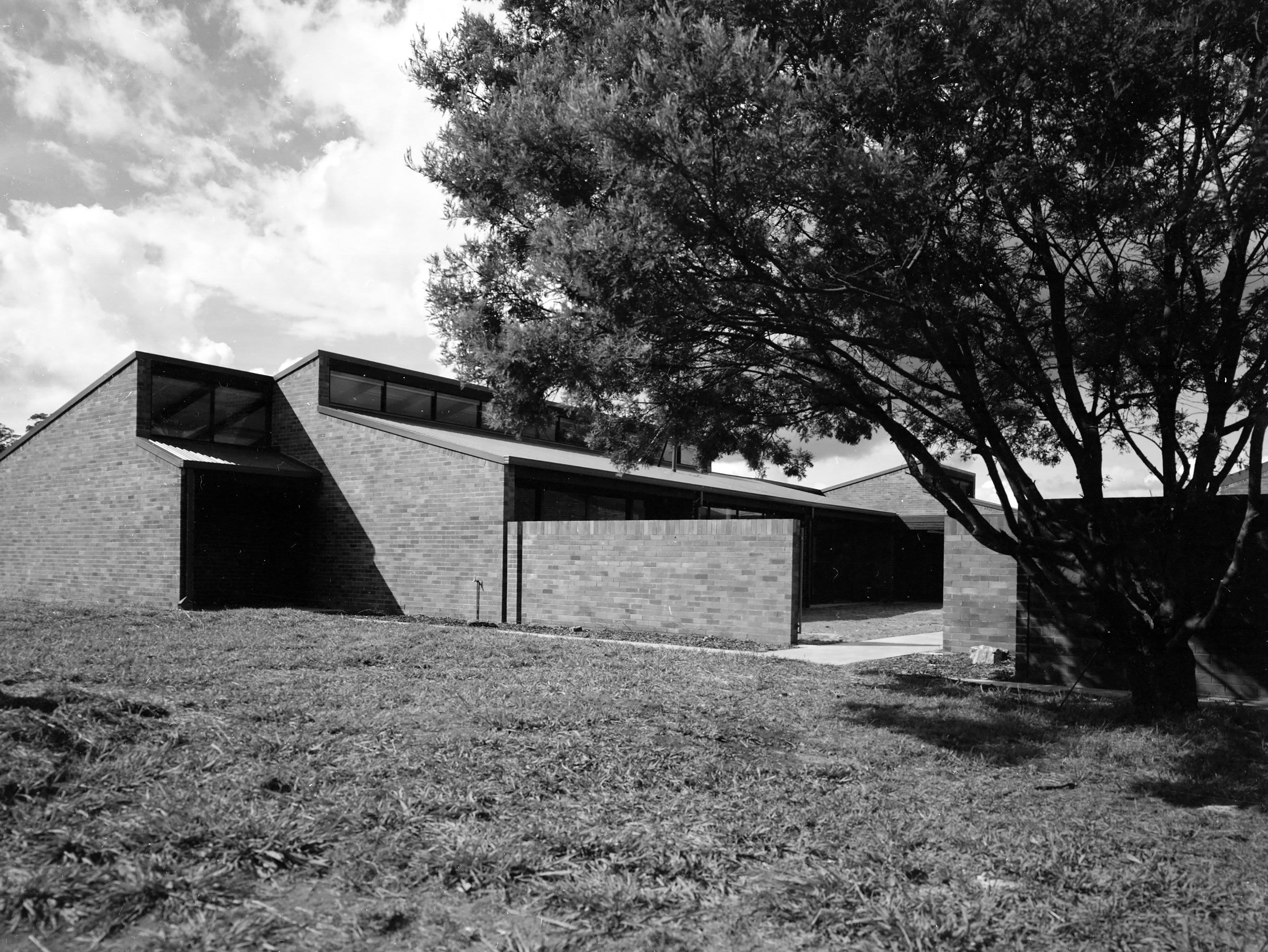 Black and white photo or a brick building with grass out the front and a big tree on the right