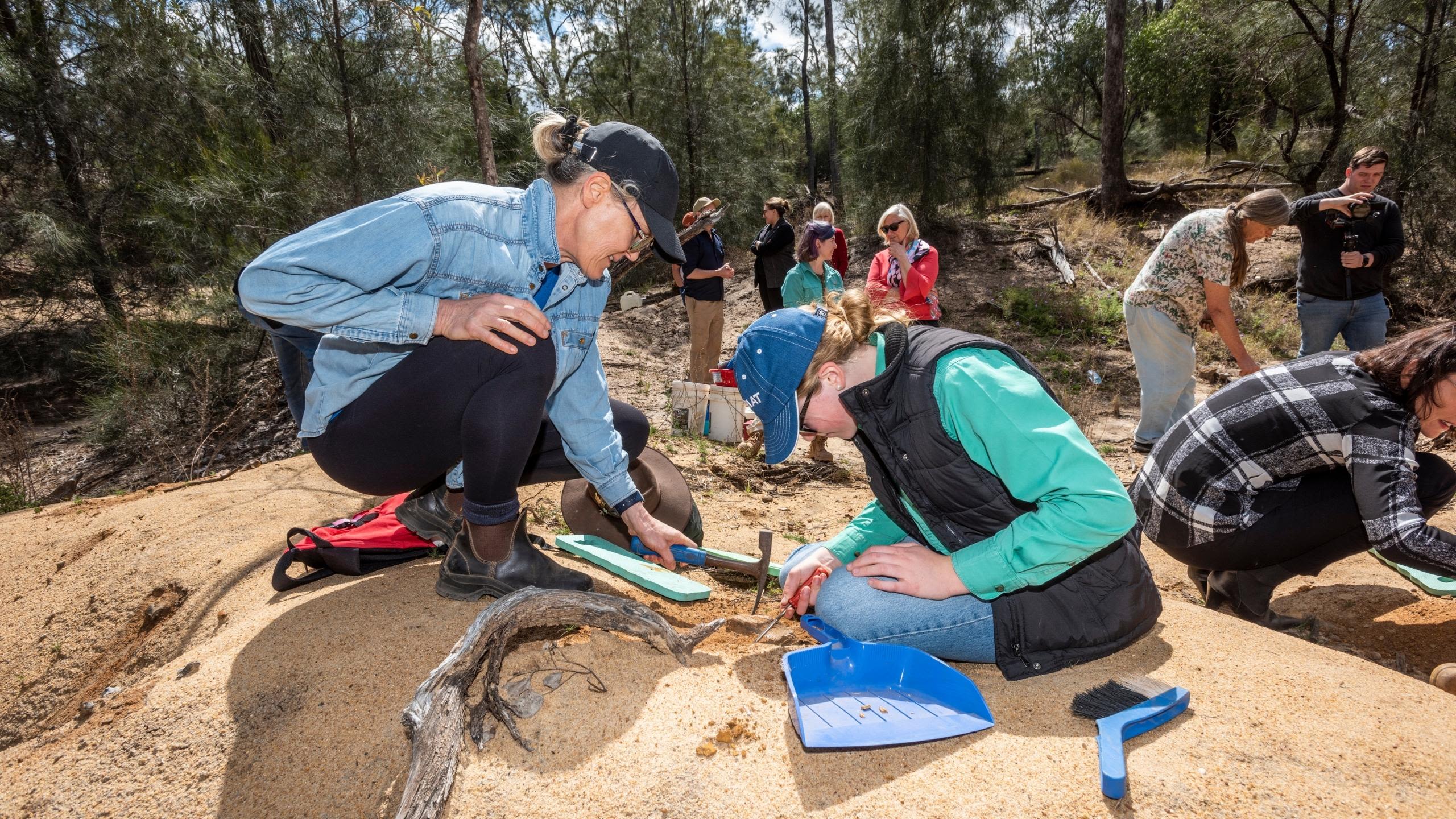 A women and a high school student sit on the sand and sift through palaeontology specimens 