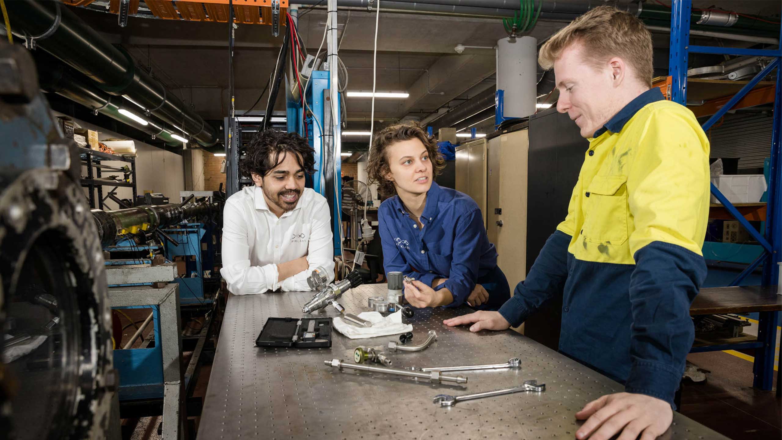 An image of Herik Labs team members (from left) Toby van den Herik, Simone Wilson and Isaiah Stook standing around a work bench looking at tools and parts.