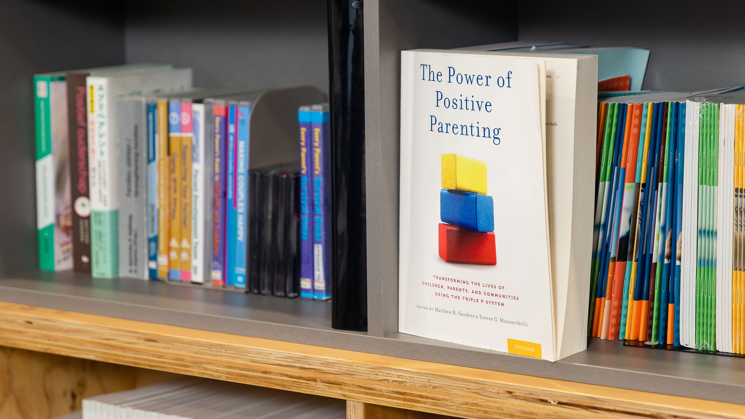 A shelf full of books. All have their spines facing outwards except for the book 'The Power of Positive Parenting' by Matthew Sanders, which has its cover facing outward.