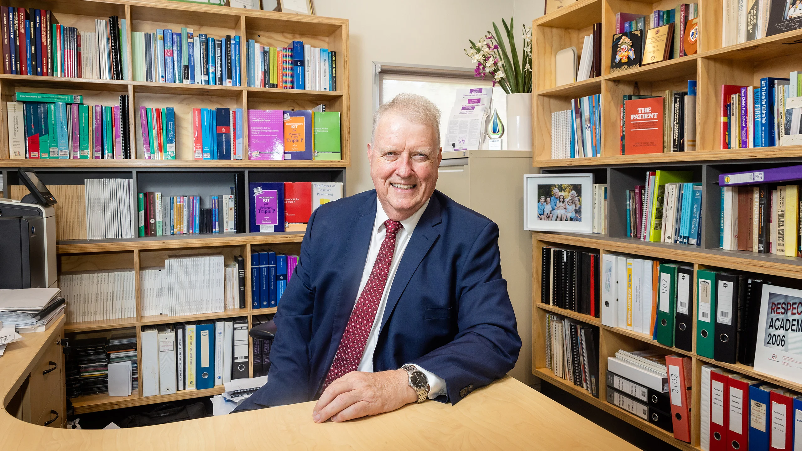 Professor Matthew Sanders, an older man, sitting and smiling at a desk in an office in front of two full bookshelves and a small filing cabinet