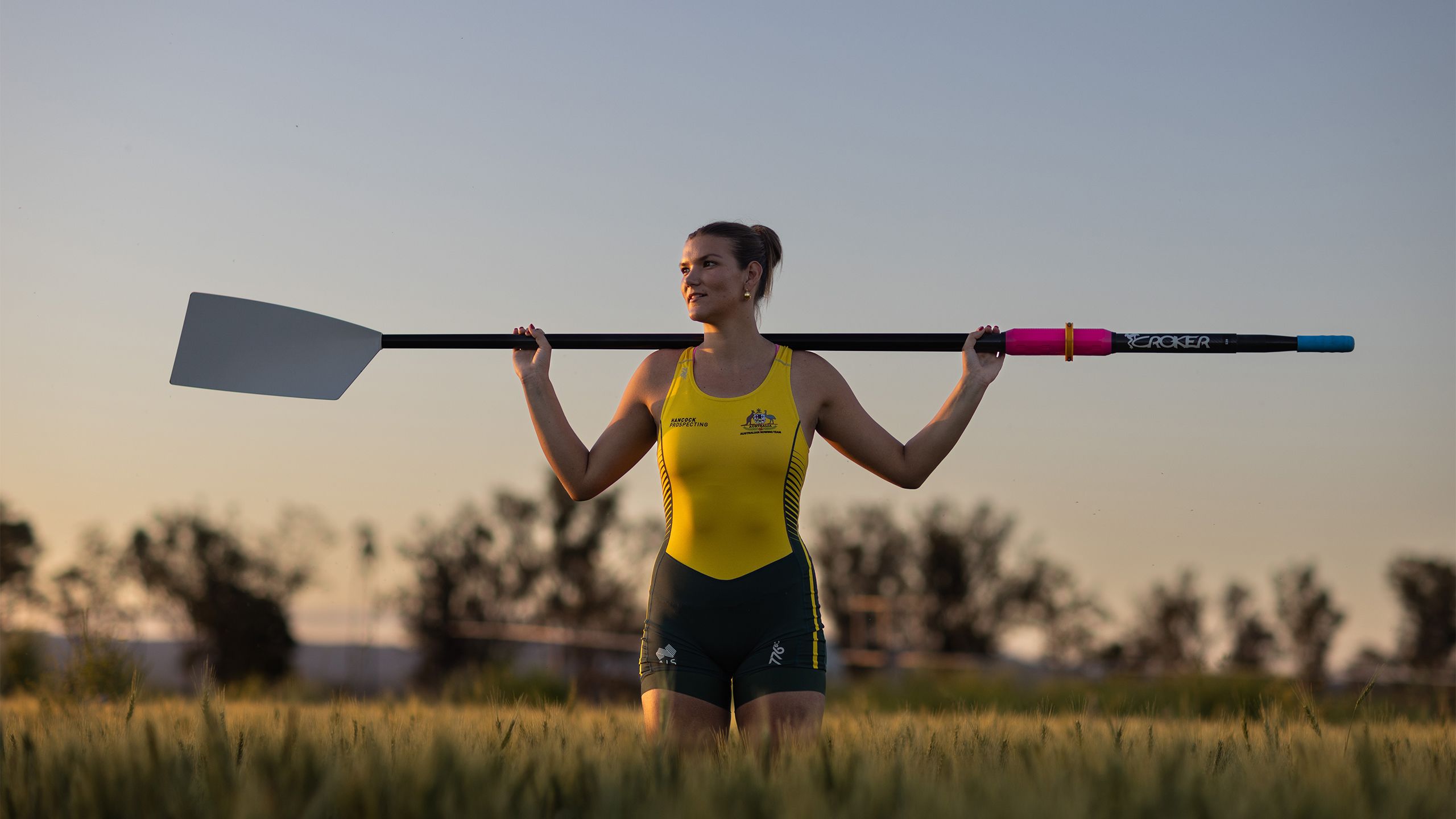 An image of UQ agribusiness student and Australian rowing representative standing in a paddock of wheat rows at UQ's Gatton campus. She is wearing her Australian rowing zoot suit and has an oar resting on both of her shoulders as the sun sets.