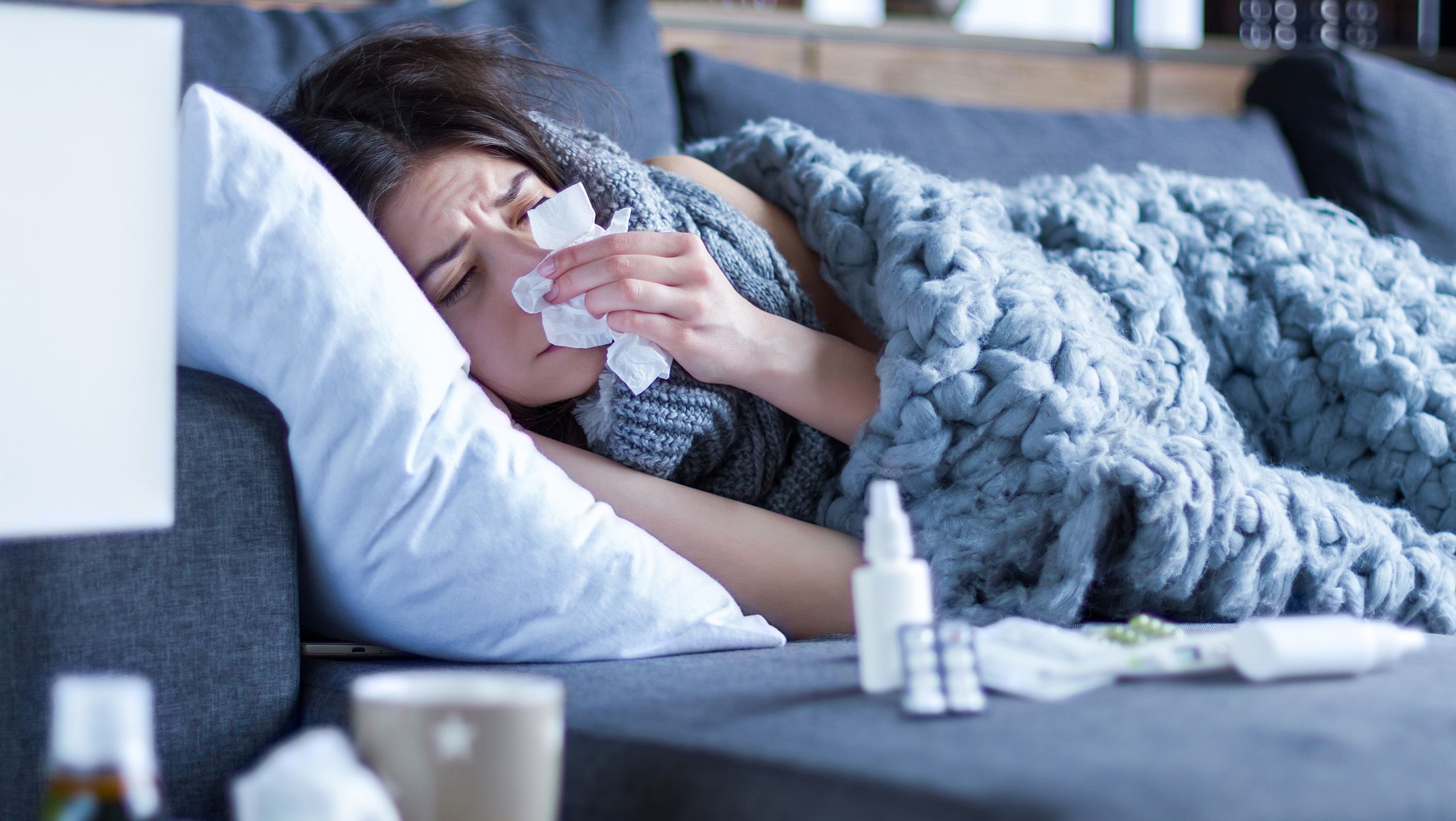 A woman lying underneath a blanket on a couch with a pillow under her head and blowing her nose
