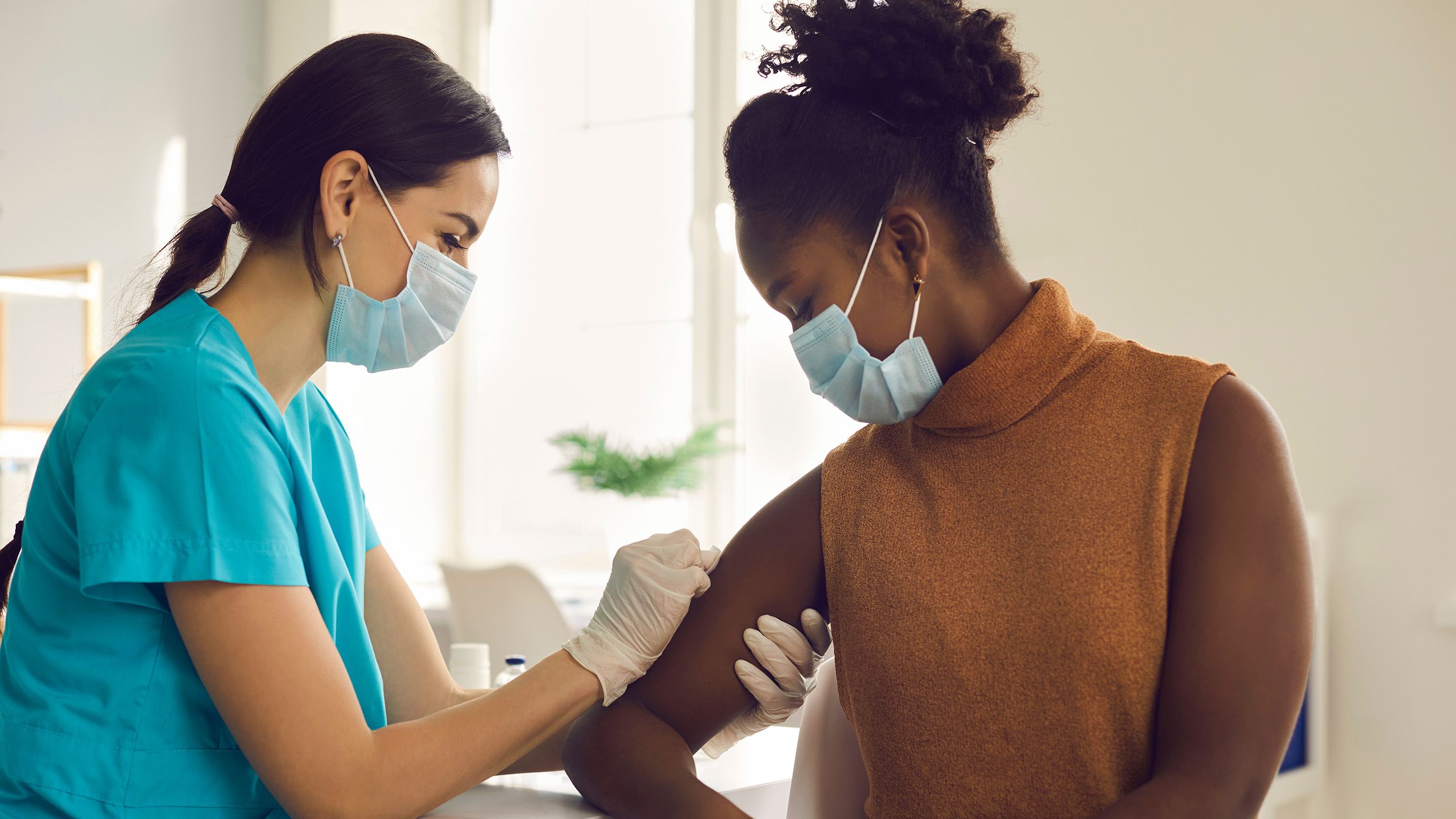 Two women wearing masks. One is preparing the other's arm to receive a vaccination