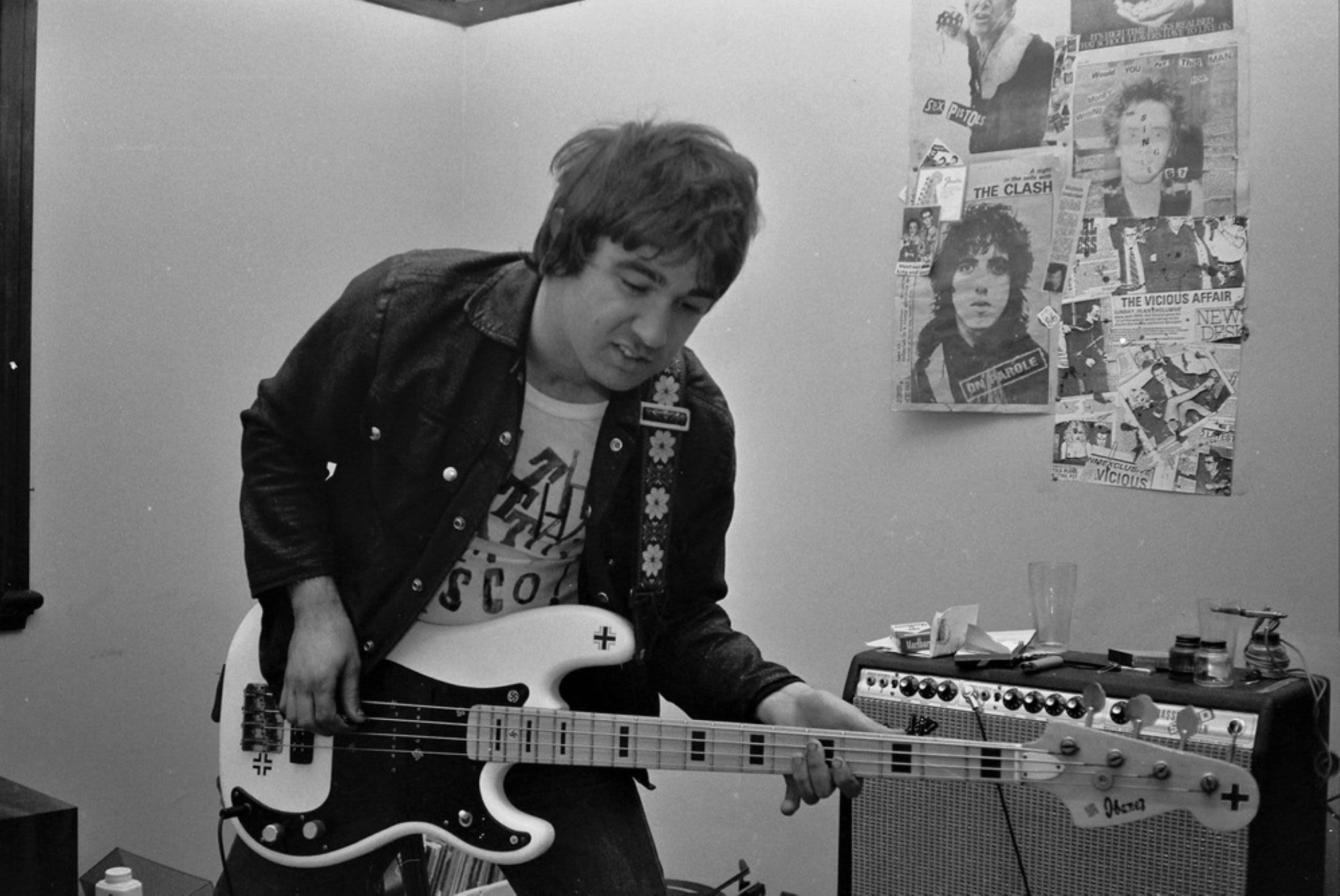 A man in leather jacket plays electric guitar with newspaper cutouts of punk music acts pinned on a wall beside him. 