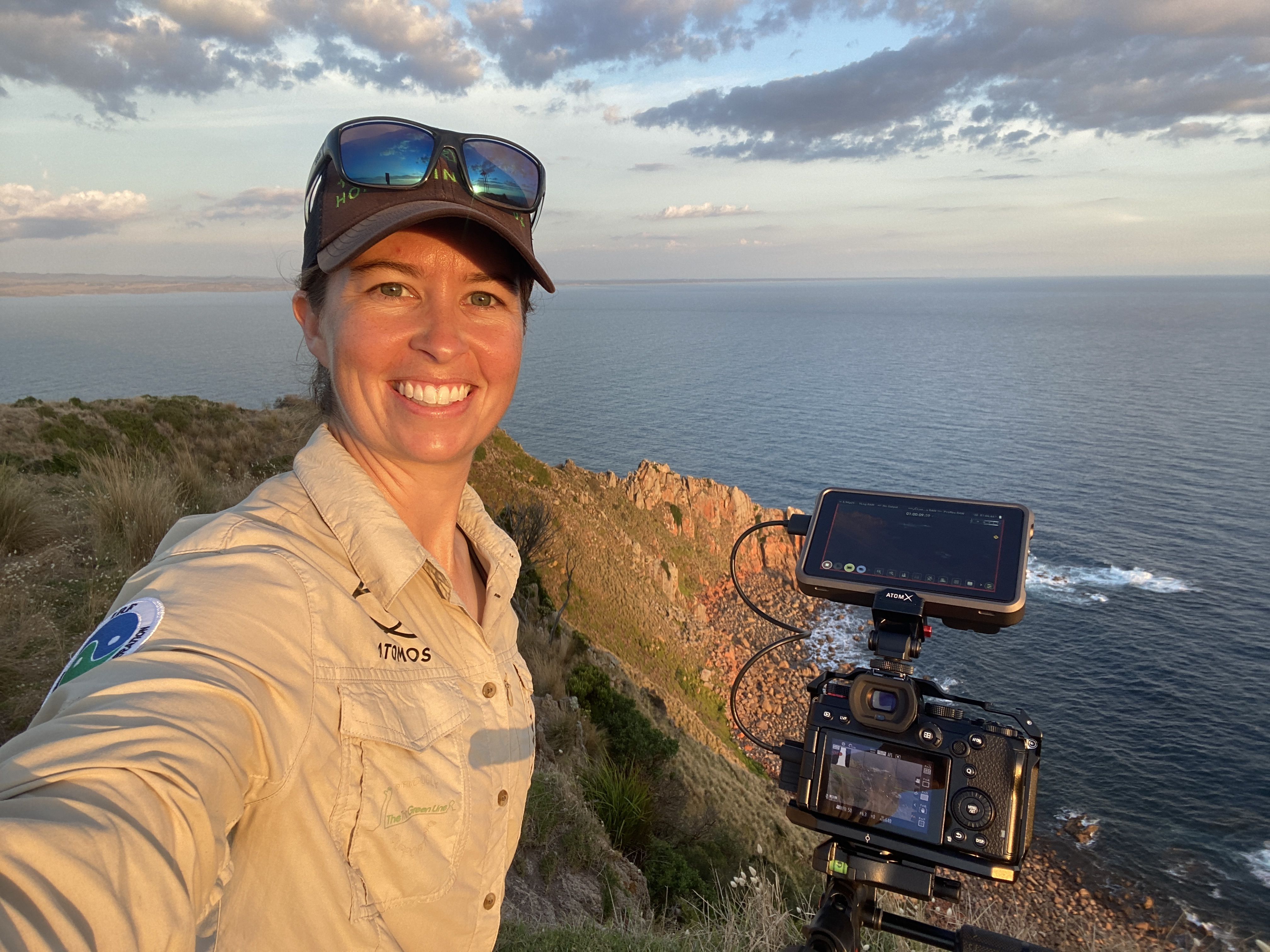 A smiling woman with a light collared shirt on, holding a camera and smiling, with the ocean and cliffs behind