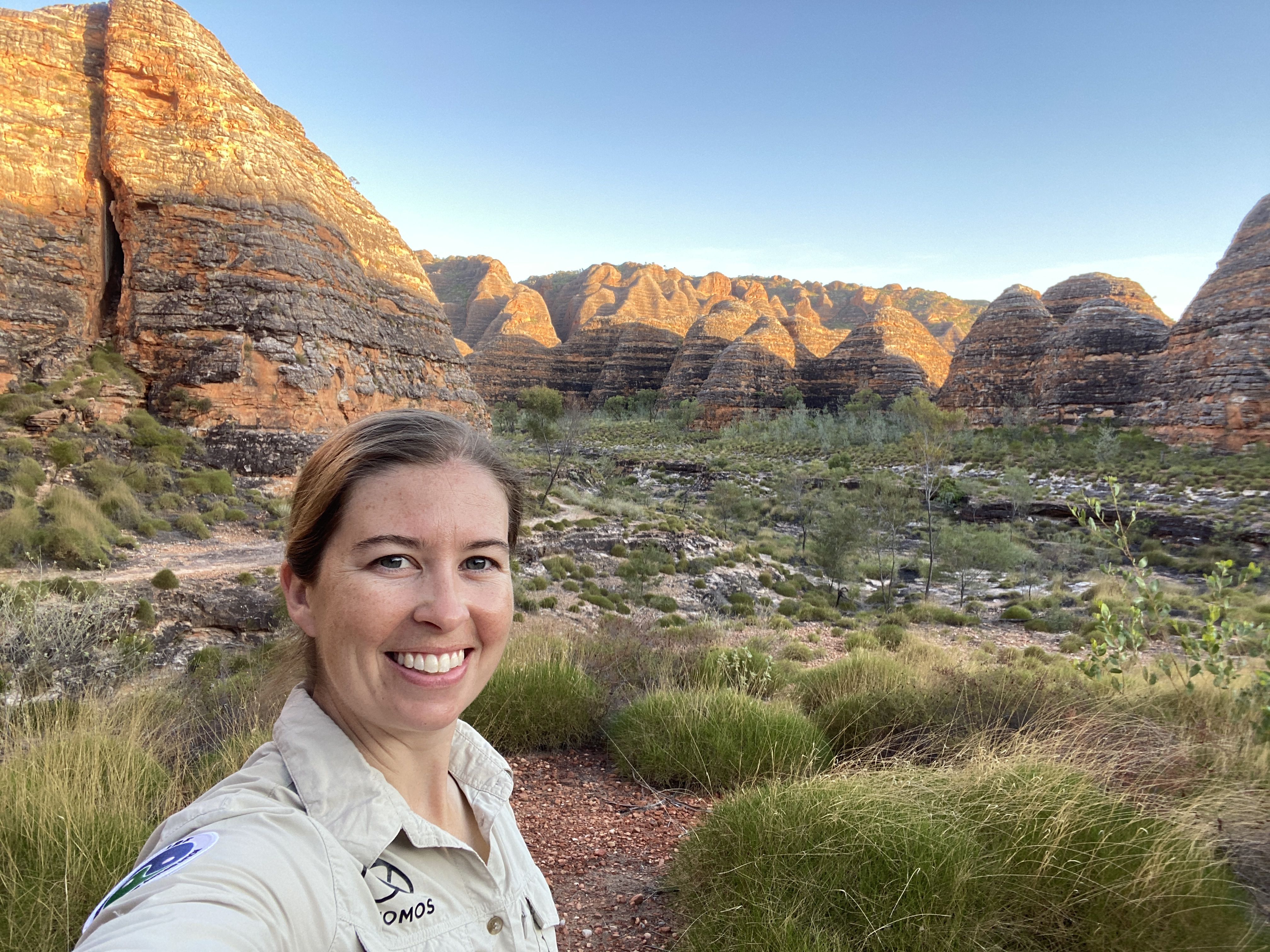 A woman smiling with red mountains and green shrubs in the background