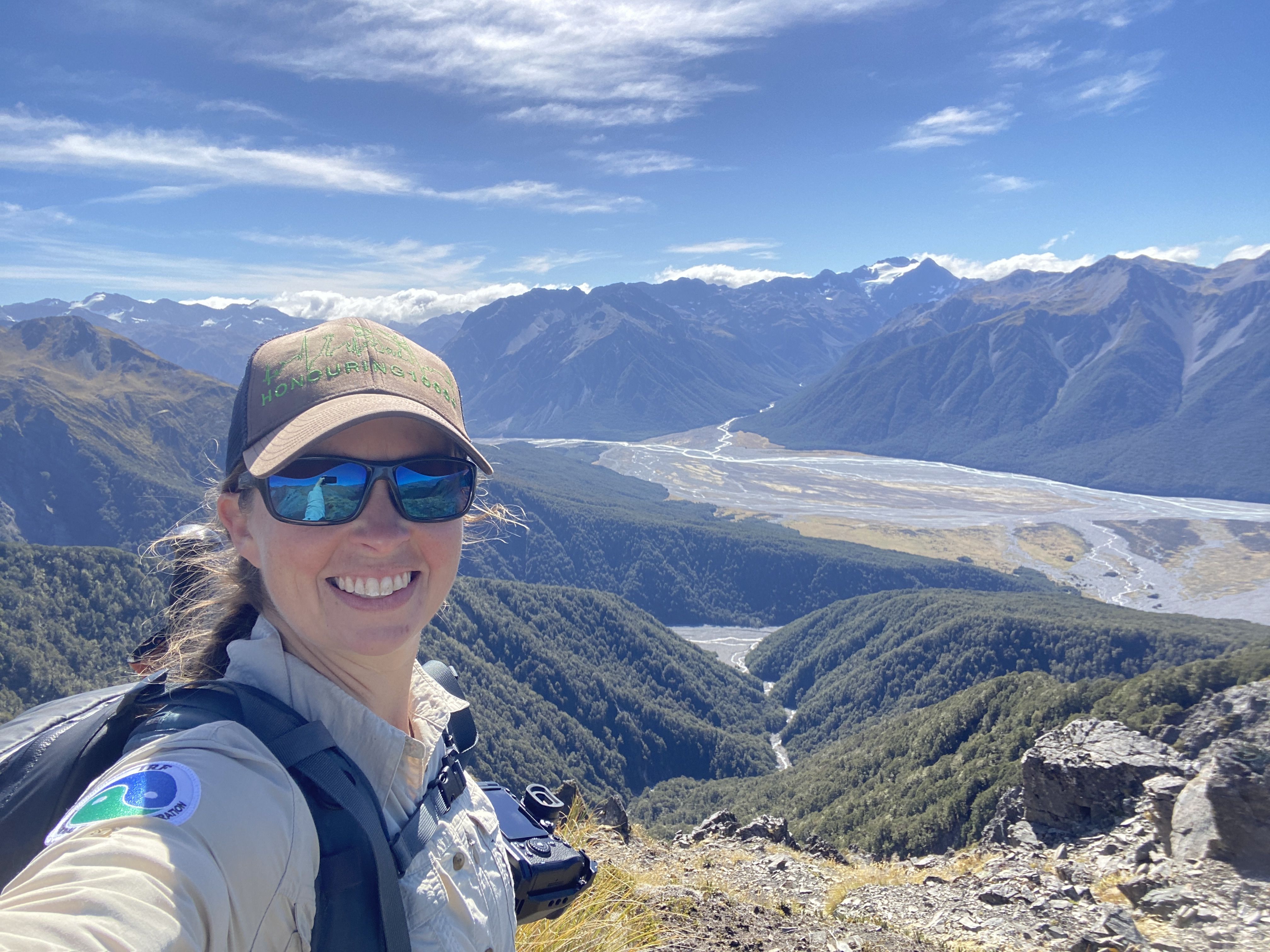 A woman's face with brown hat and blue sunglasses, with blue mountains, a blue sky, and a river behind