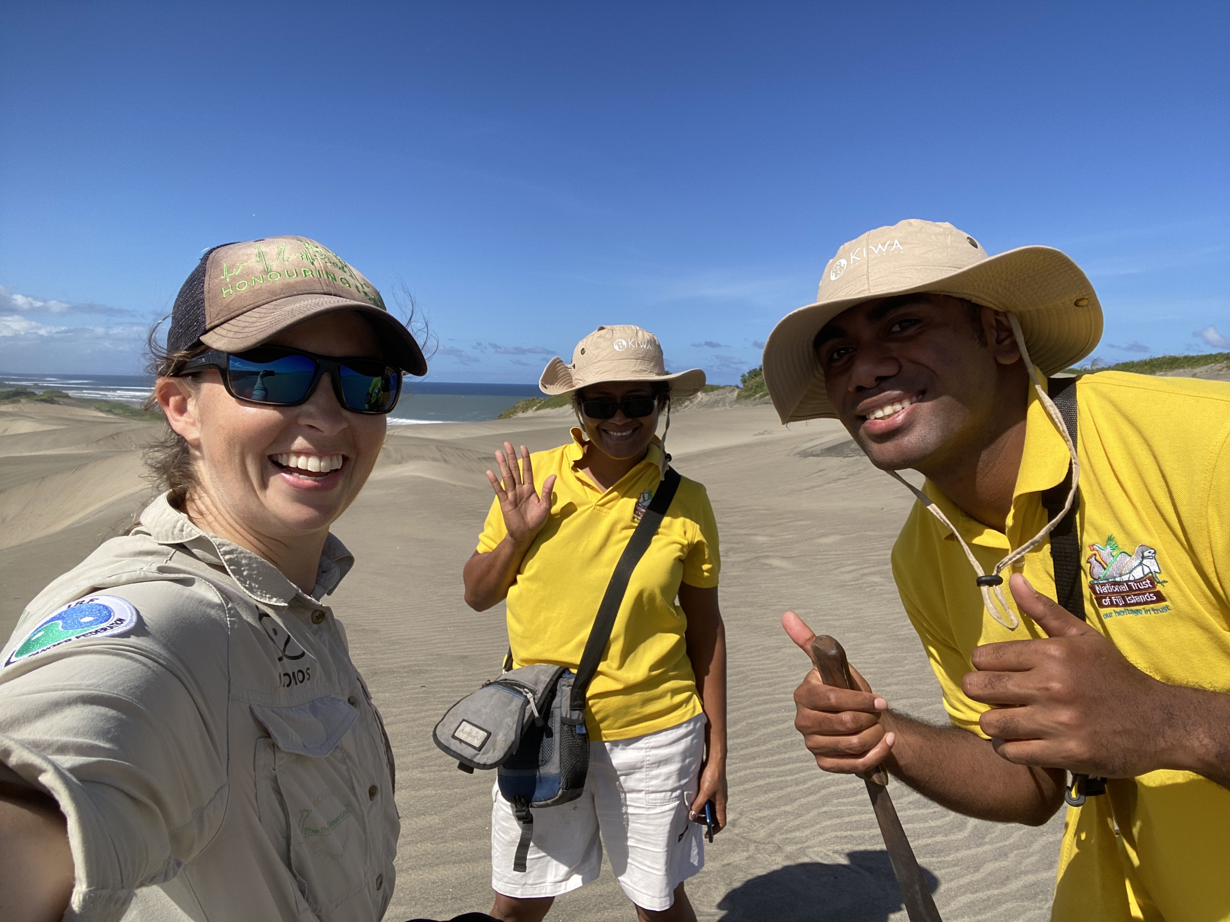 A woman with a brown hat and blue sunglasses, with a man and woman in yellow shirts and floppy hats, all smiling and giving the thumbs up