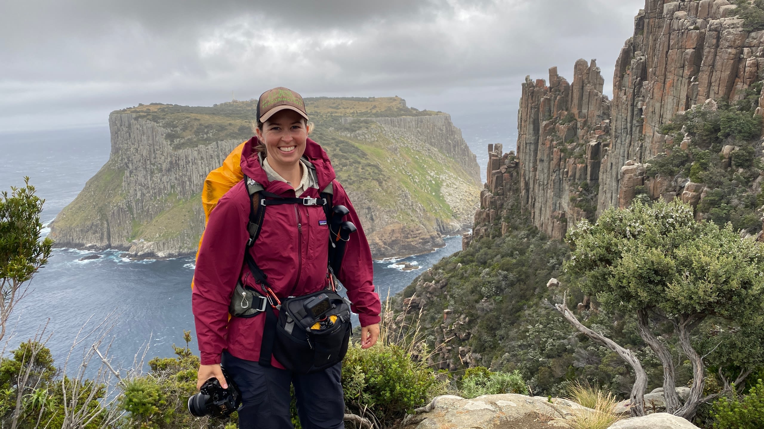 A woman in a red jacket and brown hat standing in front of the ocean and a rocky cliff with green bushes