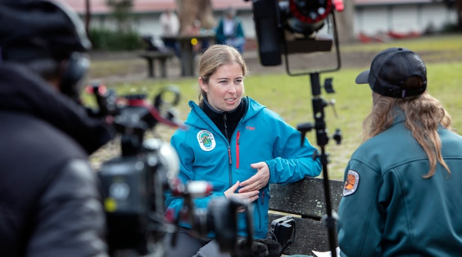 A woman in a blue jacket interviewing a woman in a green jacket, with cameras and microphones