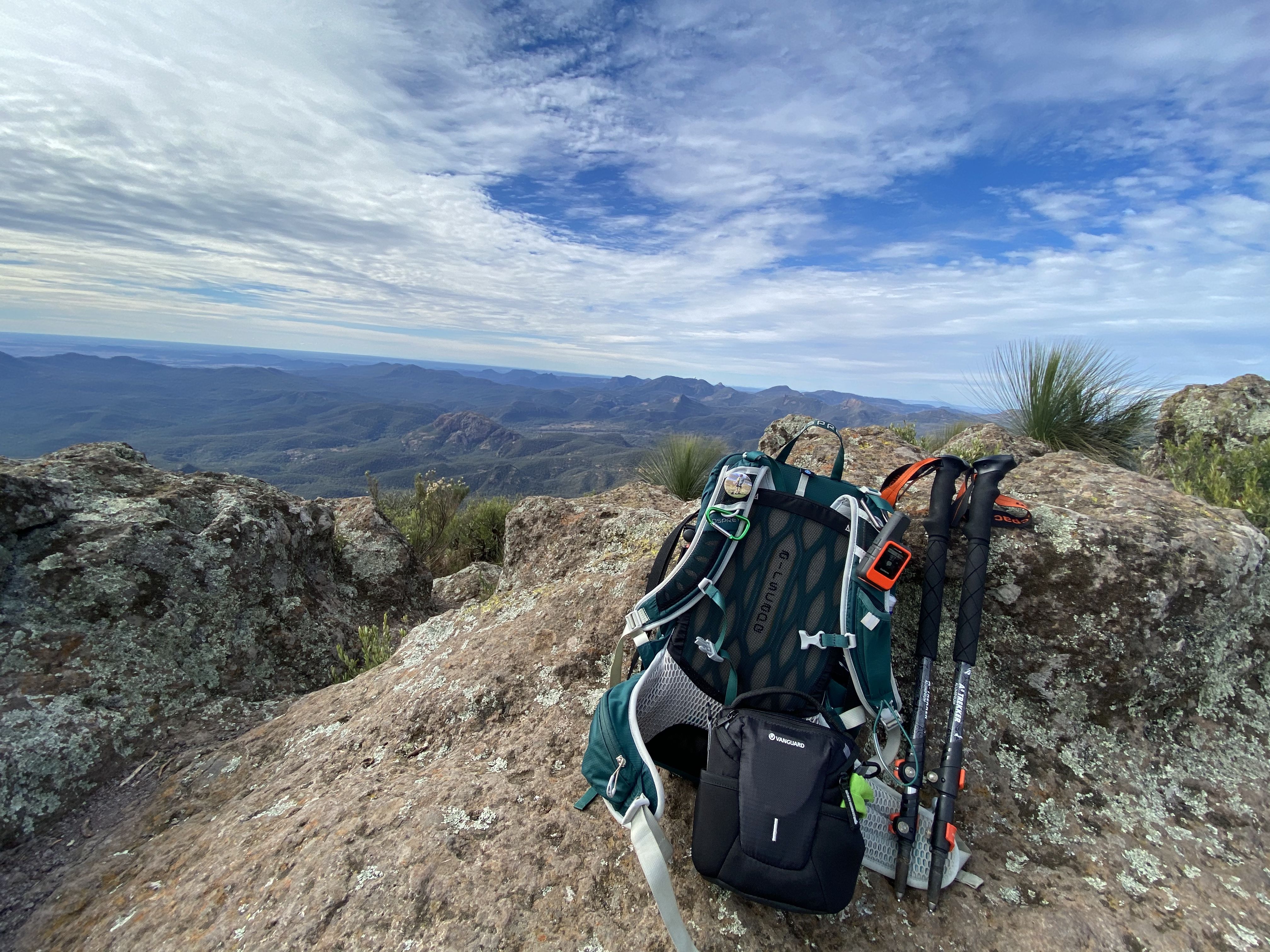 A blue backpack and orange hiking poles resting on a rock, overlooking the rolling mountains under a cloudy sky