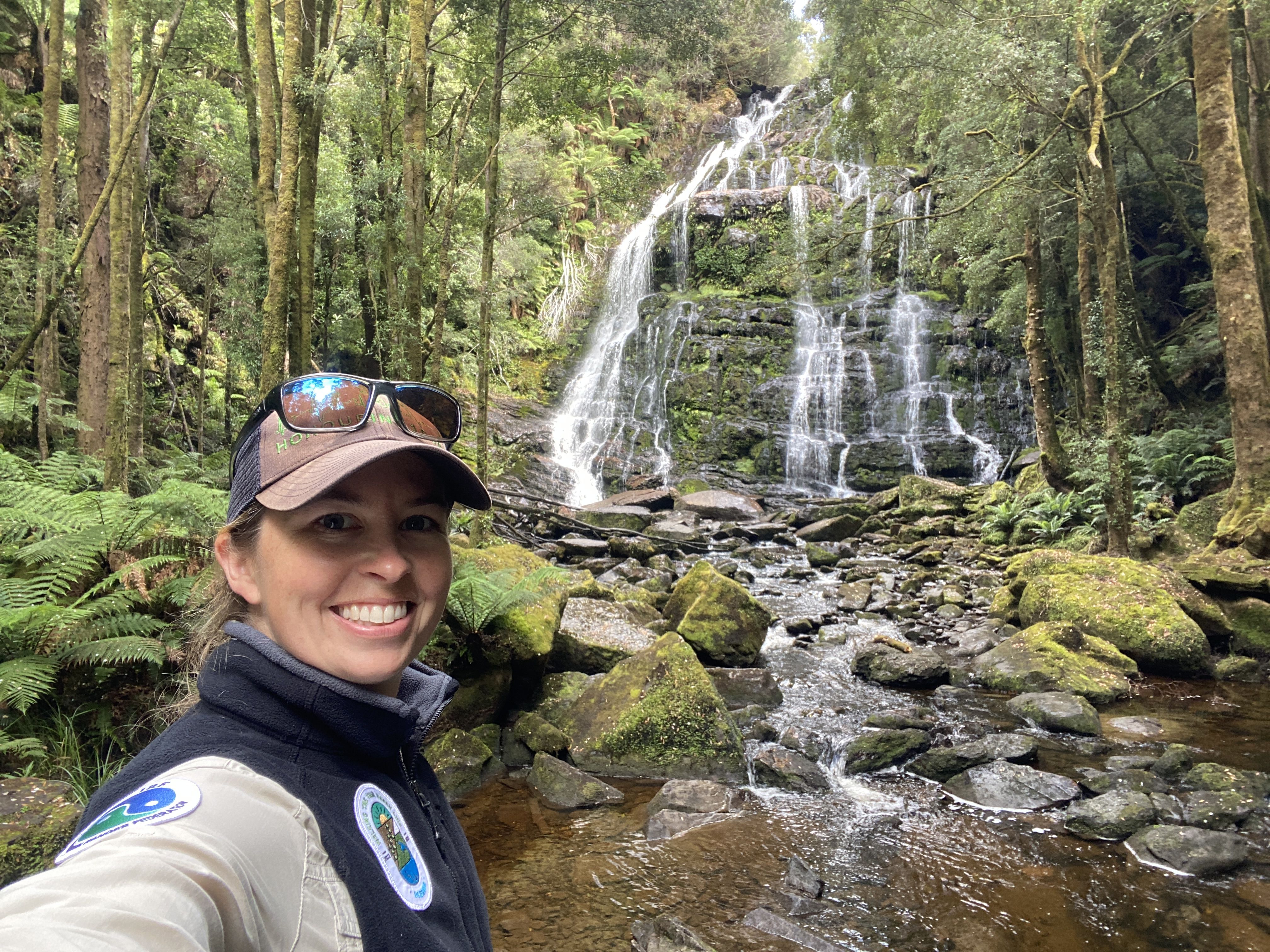 A smiling woman with a brown hat, sunglasses, and a fluffy turtleneck jumper standing in front of a cascading waterfall and mossy green rocks