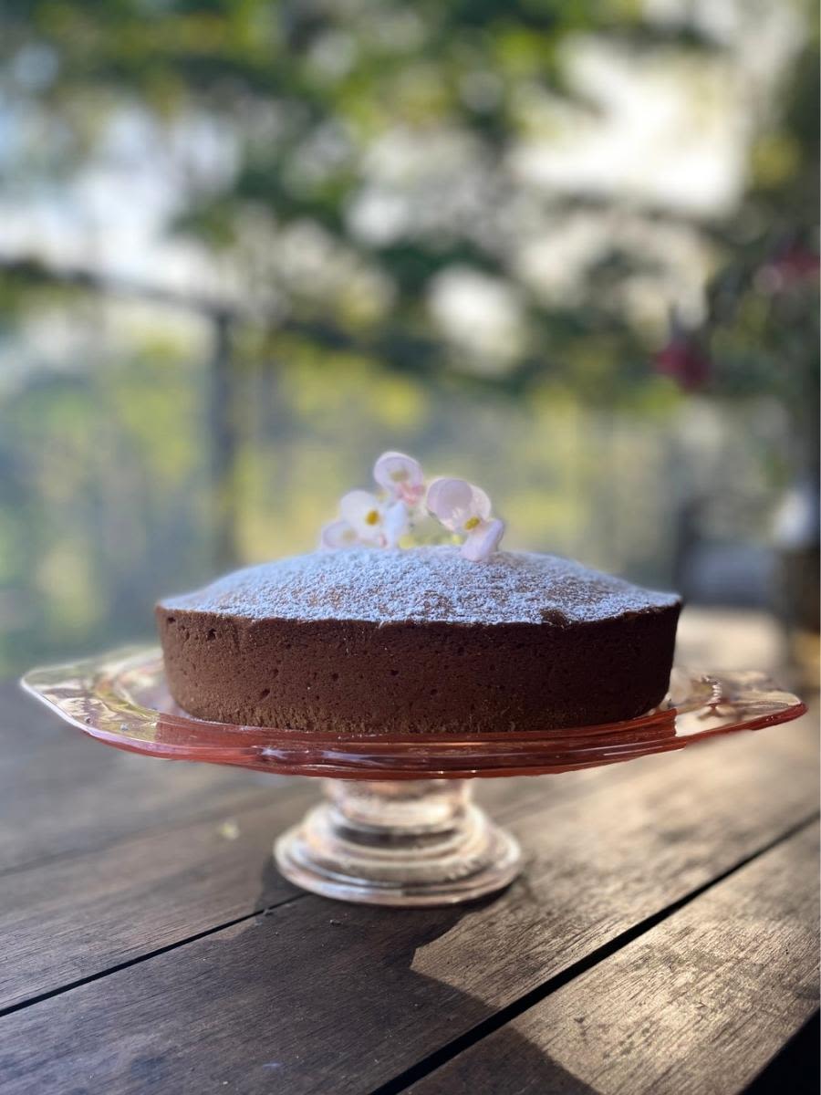 A whole brown sponge style cake dusted with icing sugar with 3 pink flowers