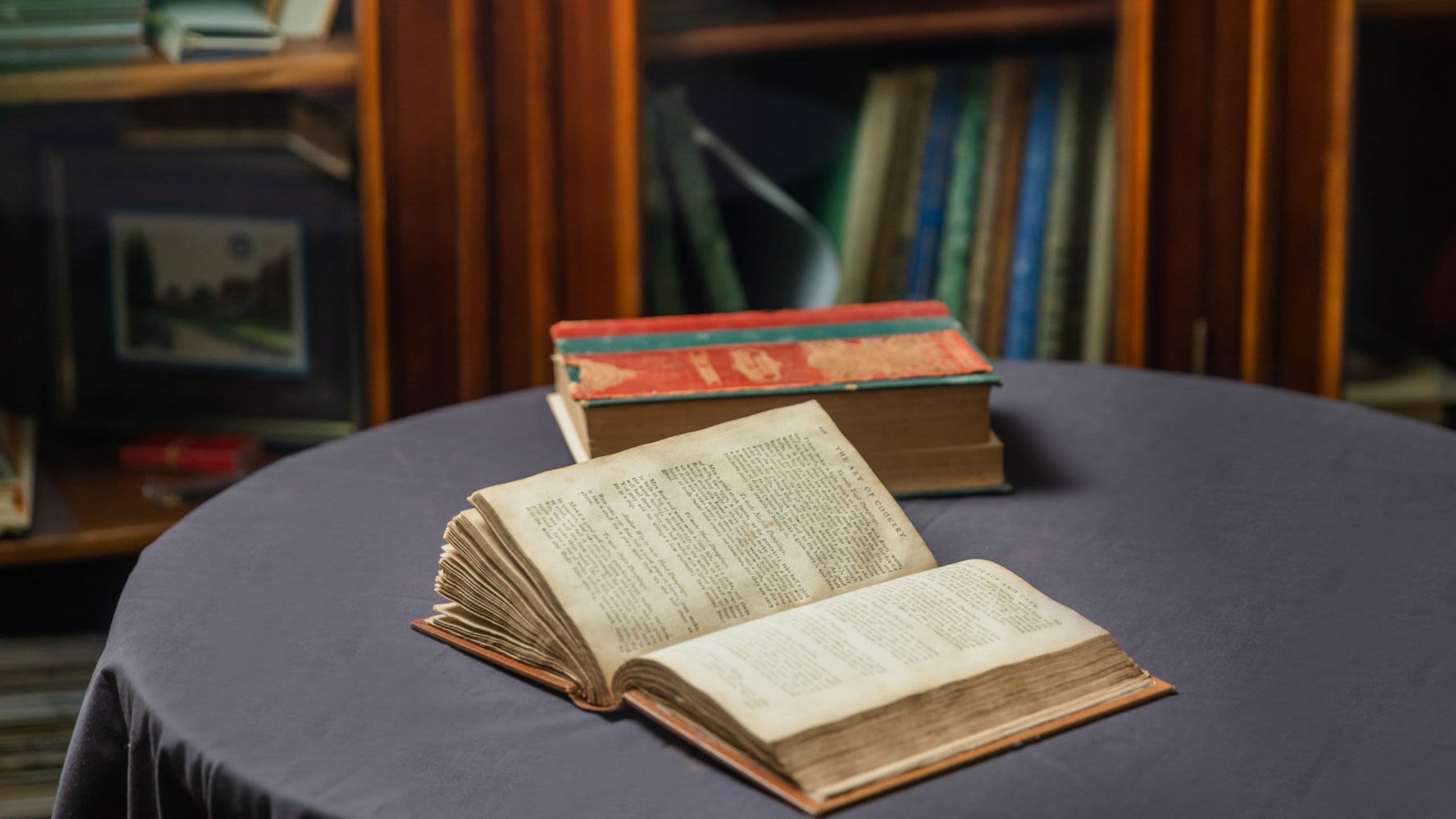 An old browned book laid open on a table with a blue tablecloth. A full bookshelf in the background