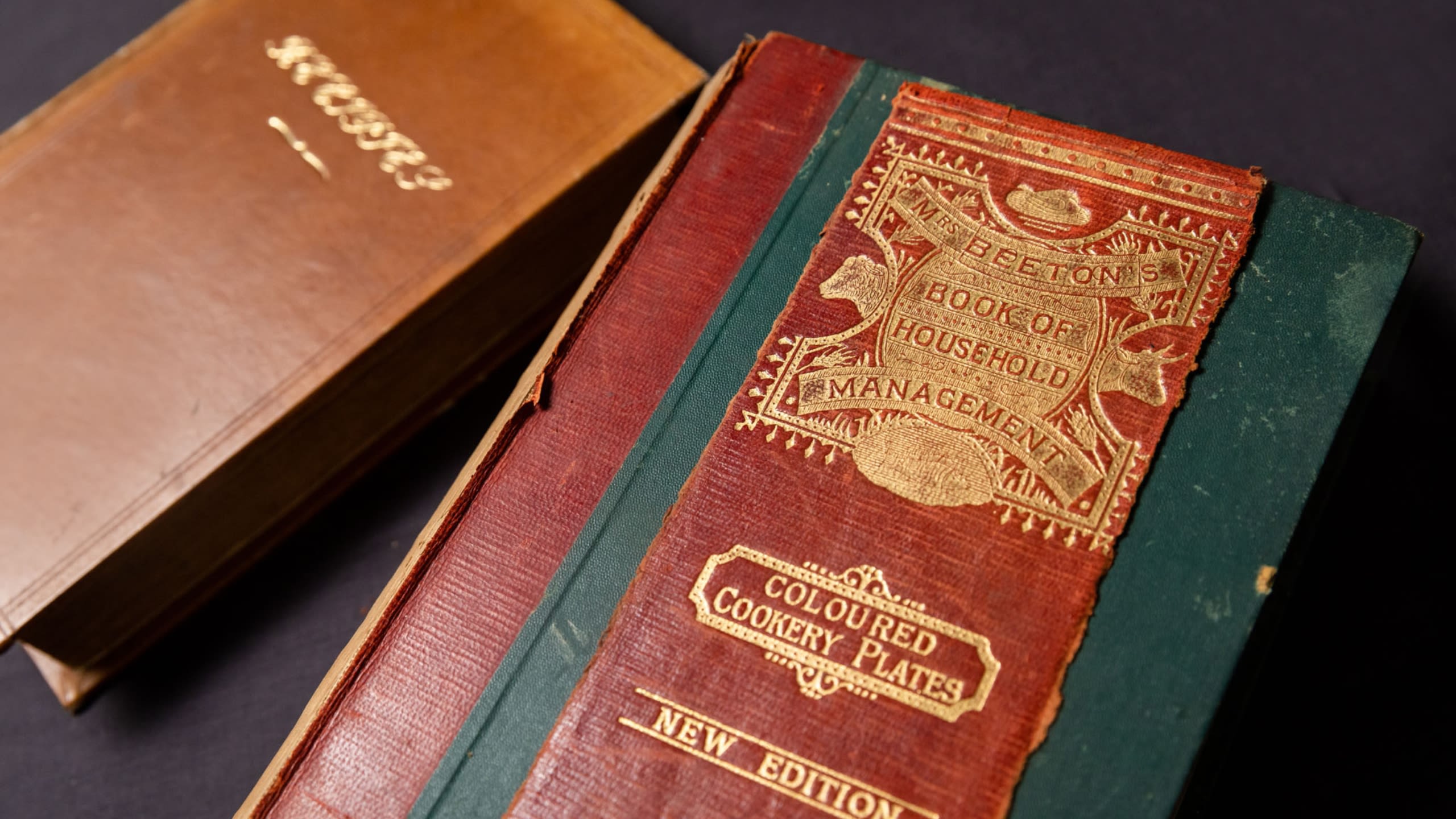 A picture of the Art of Cookery Made Plain Easy and Mrs Beeton's Book of Household Management front covers. Red and brown books with gold gilding.