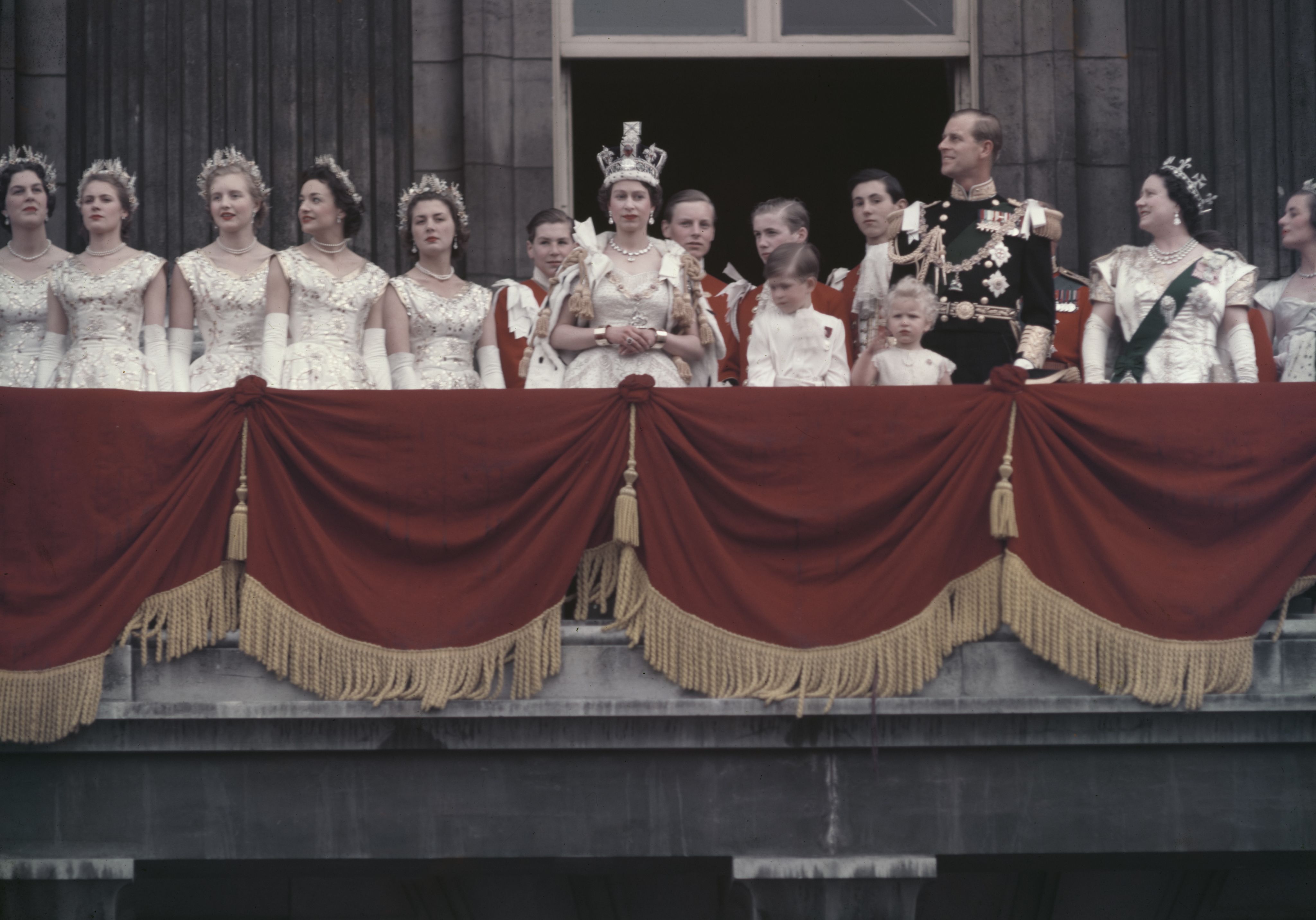 A young Queen Elizabeth standing on a balcony next to six women wearing all white, six men wearing red blazers, two young children, and Prince Philip