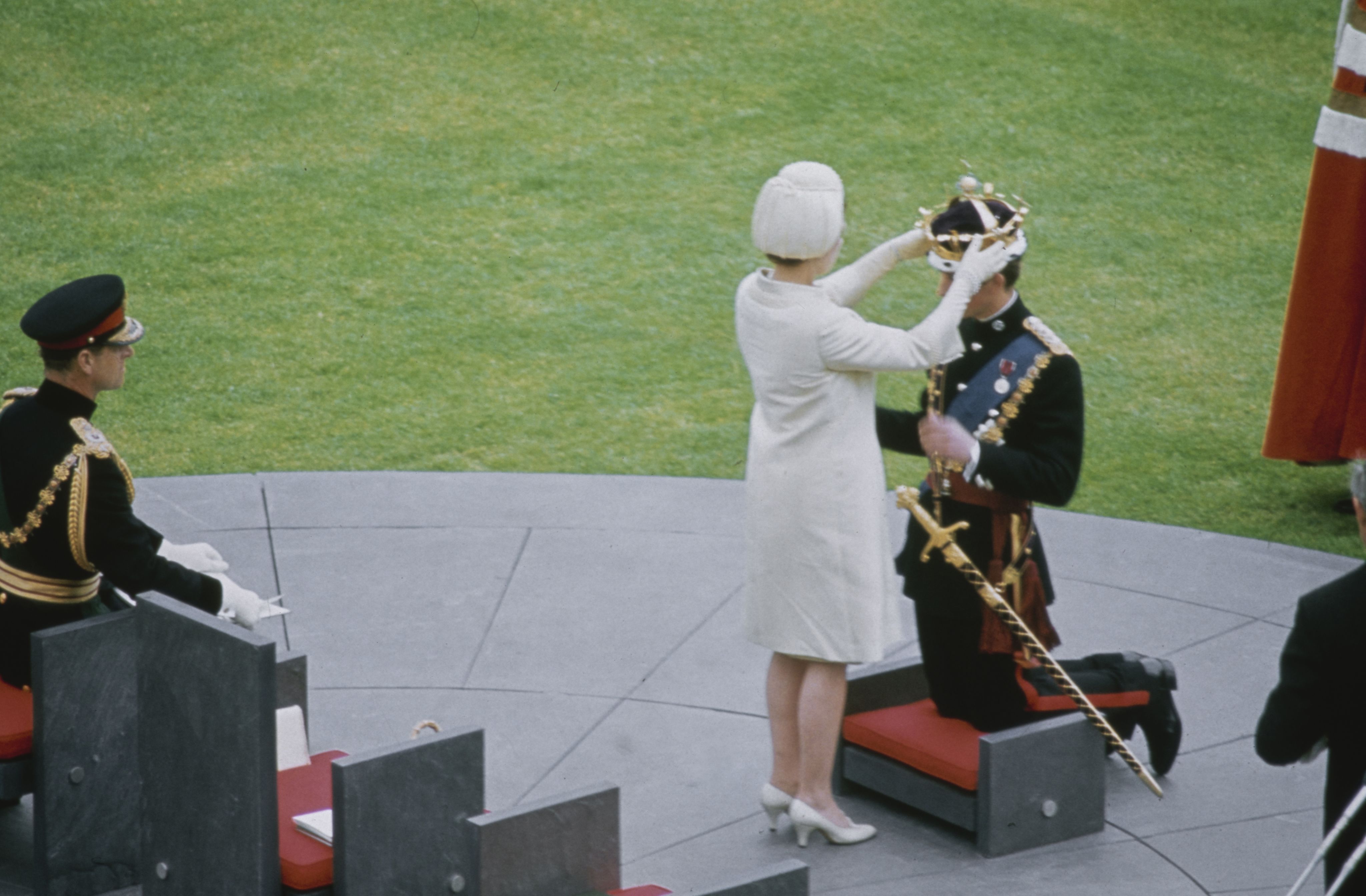The back of a woman wearing all white placing a crown on the head of a man wearing royal regalia who is kneeling on a red pedestal