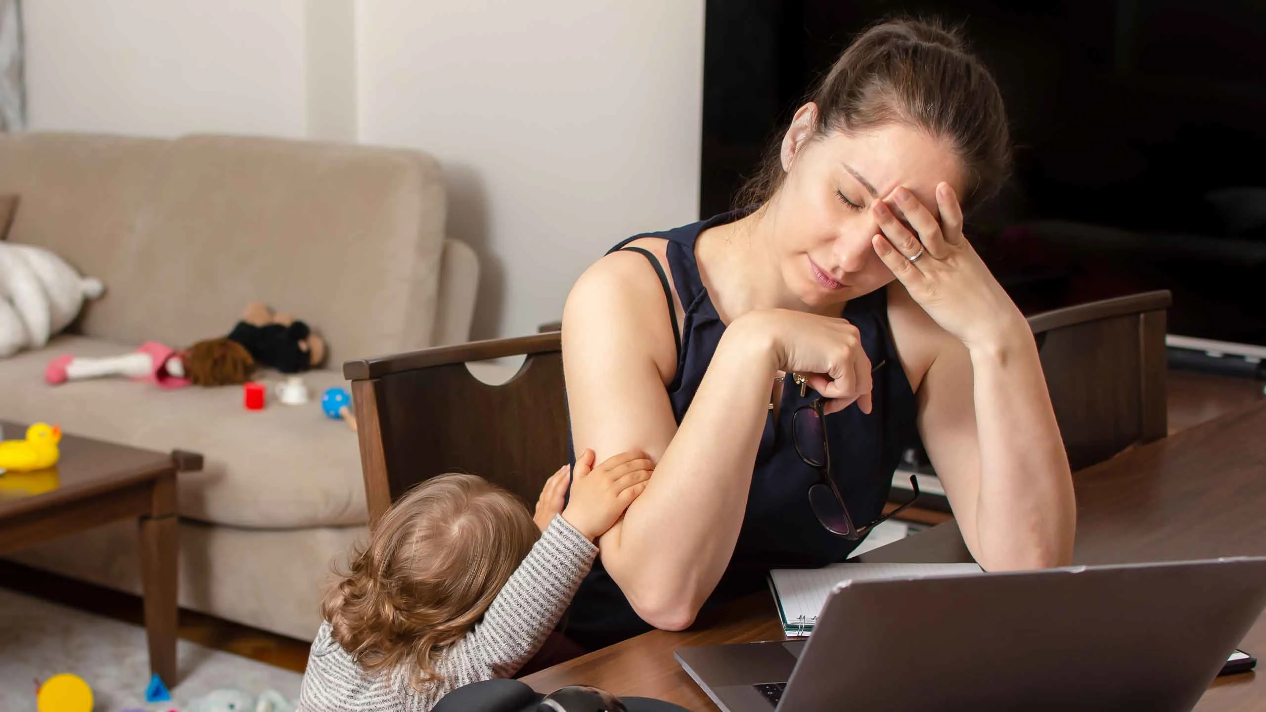 Tired mother trying to work on a laptop at home while her kid cries and holds onto her arm.