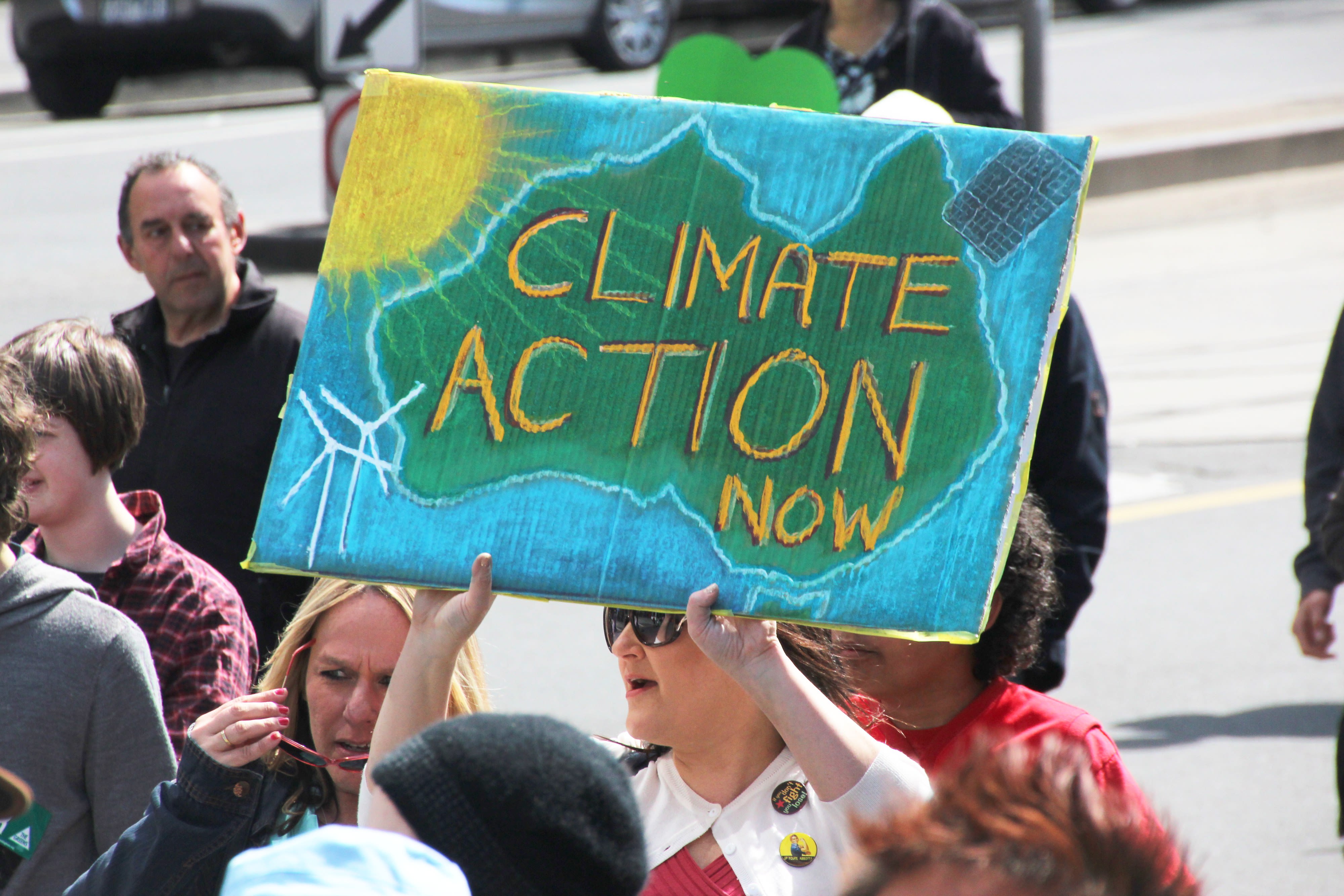 A female protester wearing sunglasses and a white long sleeve shirt shouts as she holds a protest sign. The sign portrays Australia in green painted on a sky blue background with "CLIMATE ACTION NOW" written with yellow and black crayon.