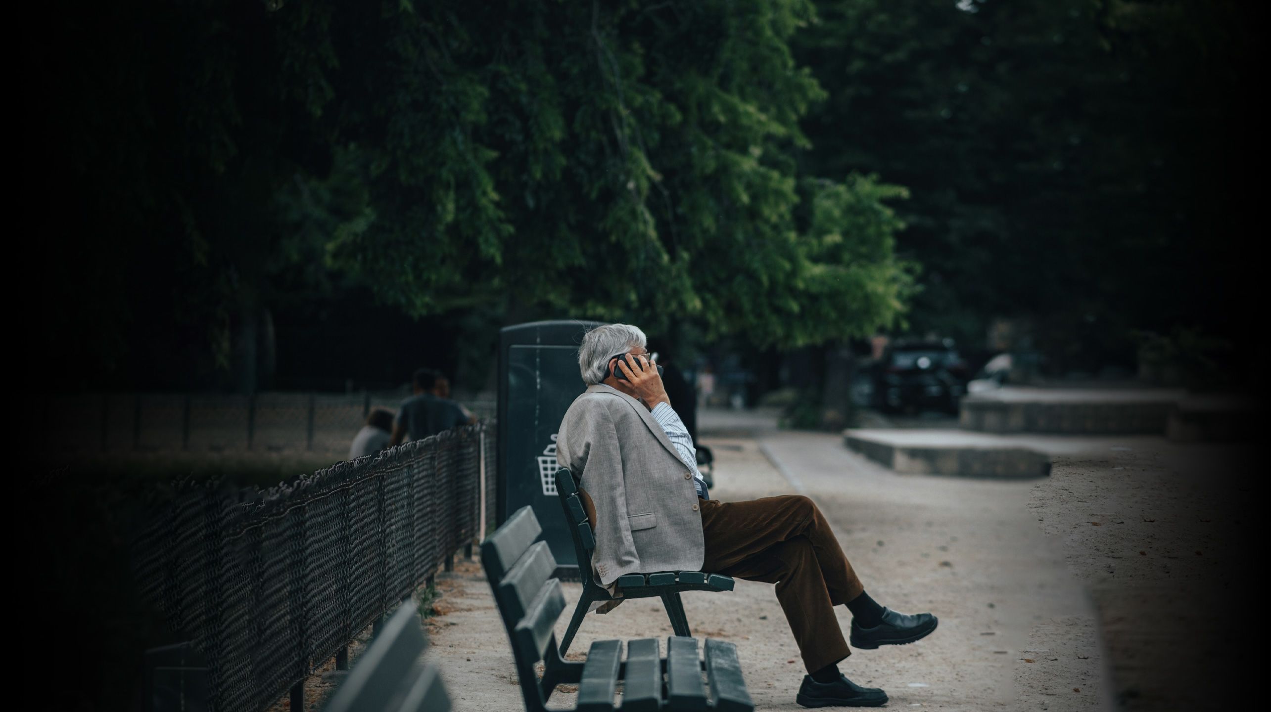 Older man sitting on a park bench, he is on the phone, he has a grey suit jacket slung over his shoulder. 
