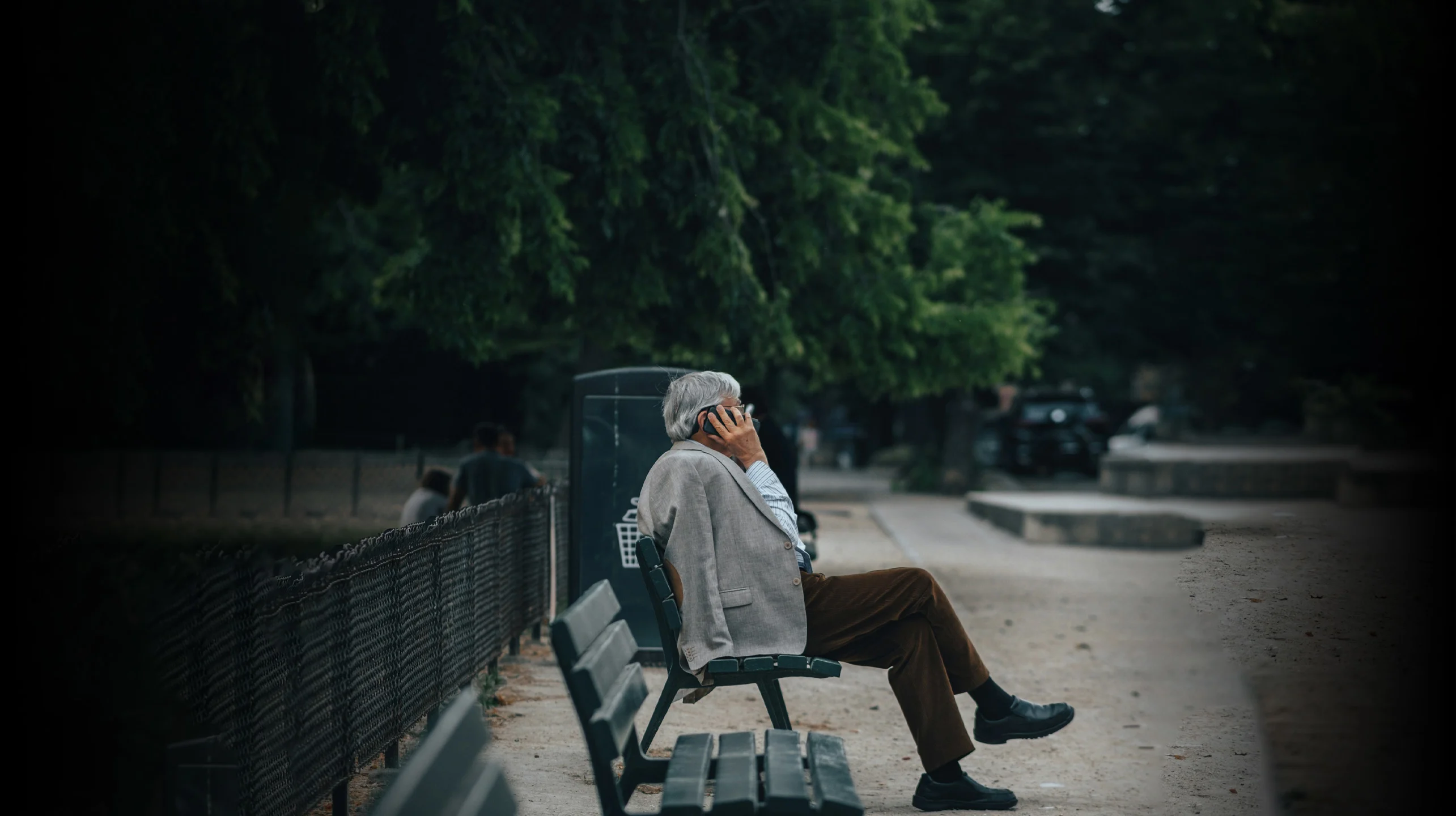 Older man sitting on a park bench, he is on the phone, he has a grey suit jacket slung over his shoulder. 
