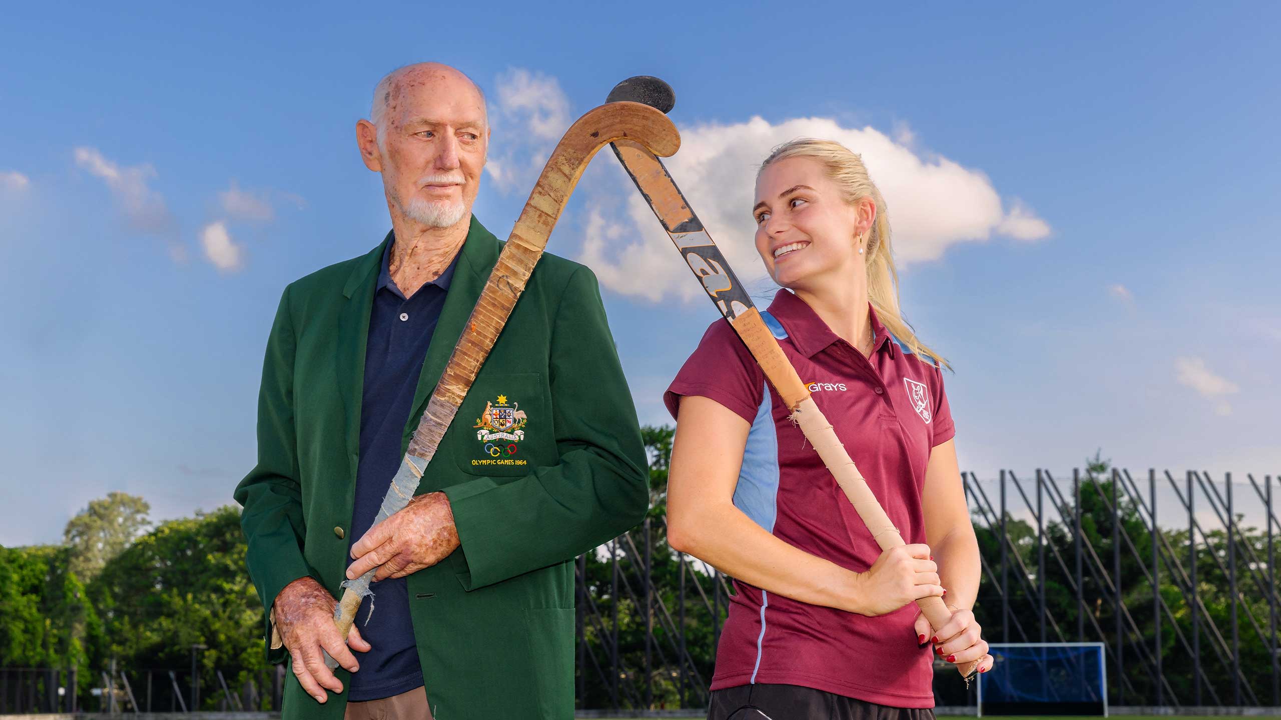 An image of UQ Hockey Club legend and dual Olympian Don McWatters and UQ Division 1 women's captain Morgan Gallagher back-to-back on the UQ hockey playing fields at UQ's St Lucia campus.