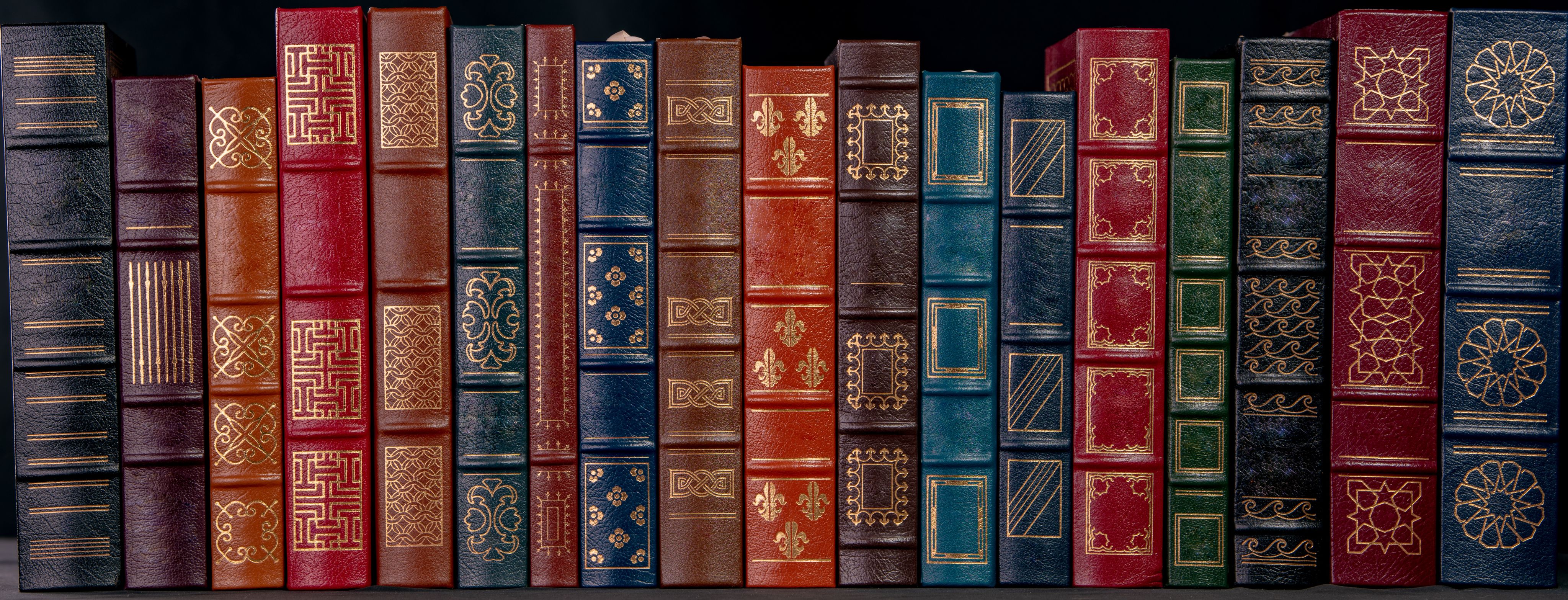 A collection of old, leather bound books in various colours sit atop a shelf.