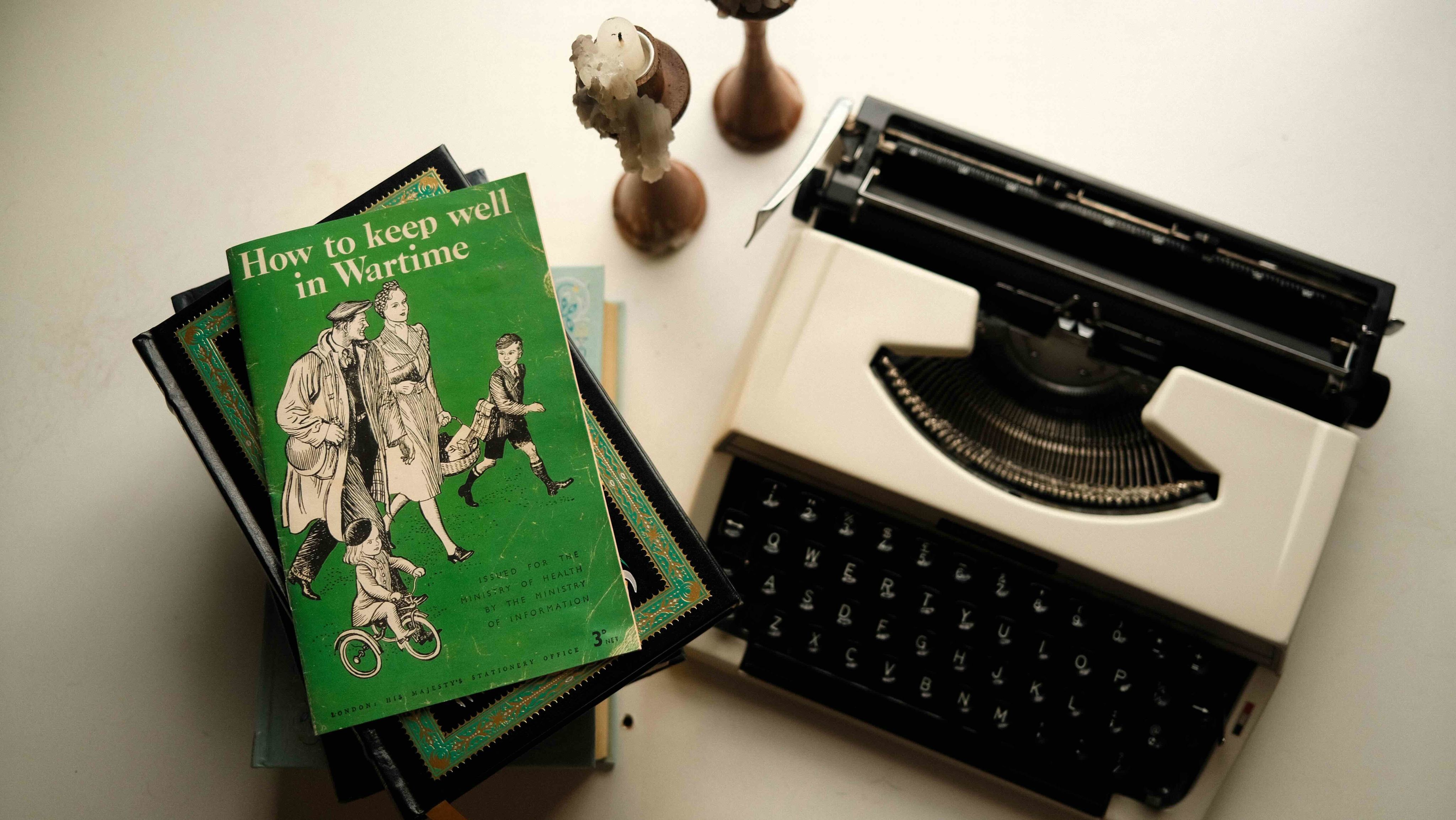An old book titled 'How to keep well in Wartime' sits beside a vintage typewriter