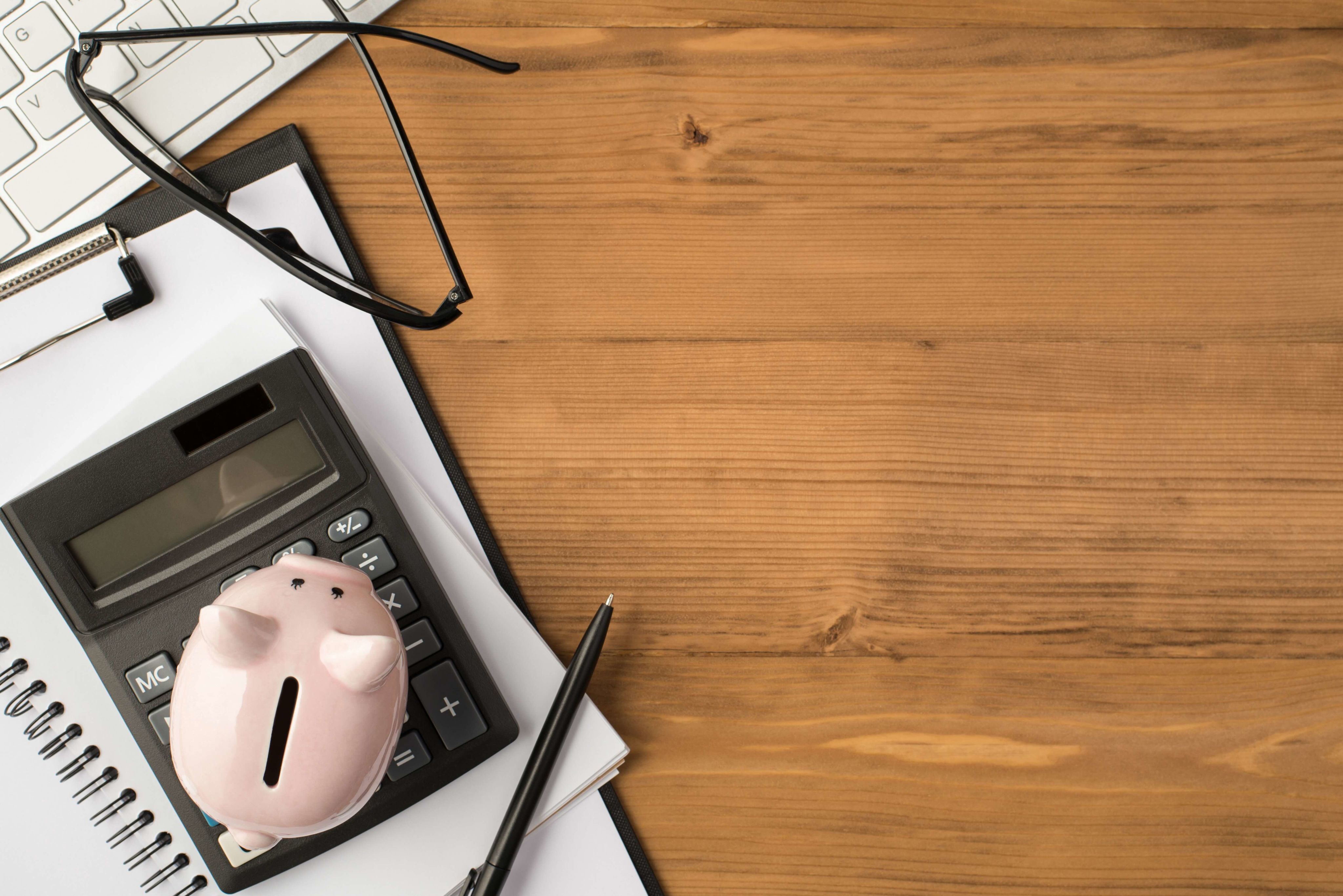 A top view image of a notepad, calculator, piggy bank and reading glasses on a dark wooden tabletop.