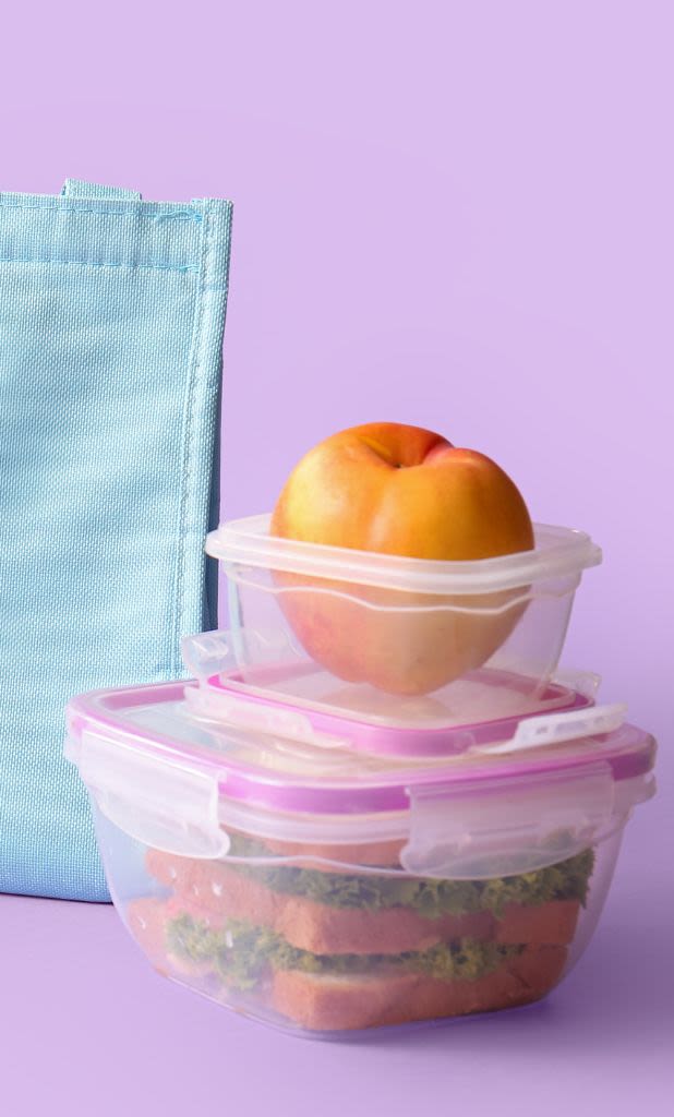 A stack of food storage containers with fruit and sandwiches against a lilac background.