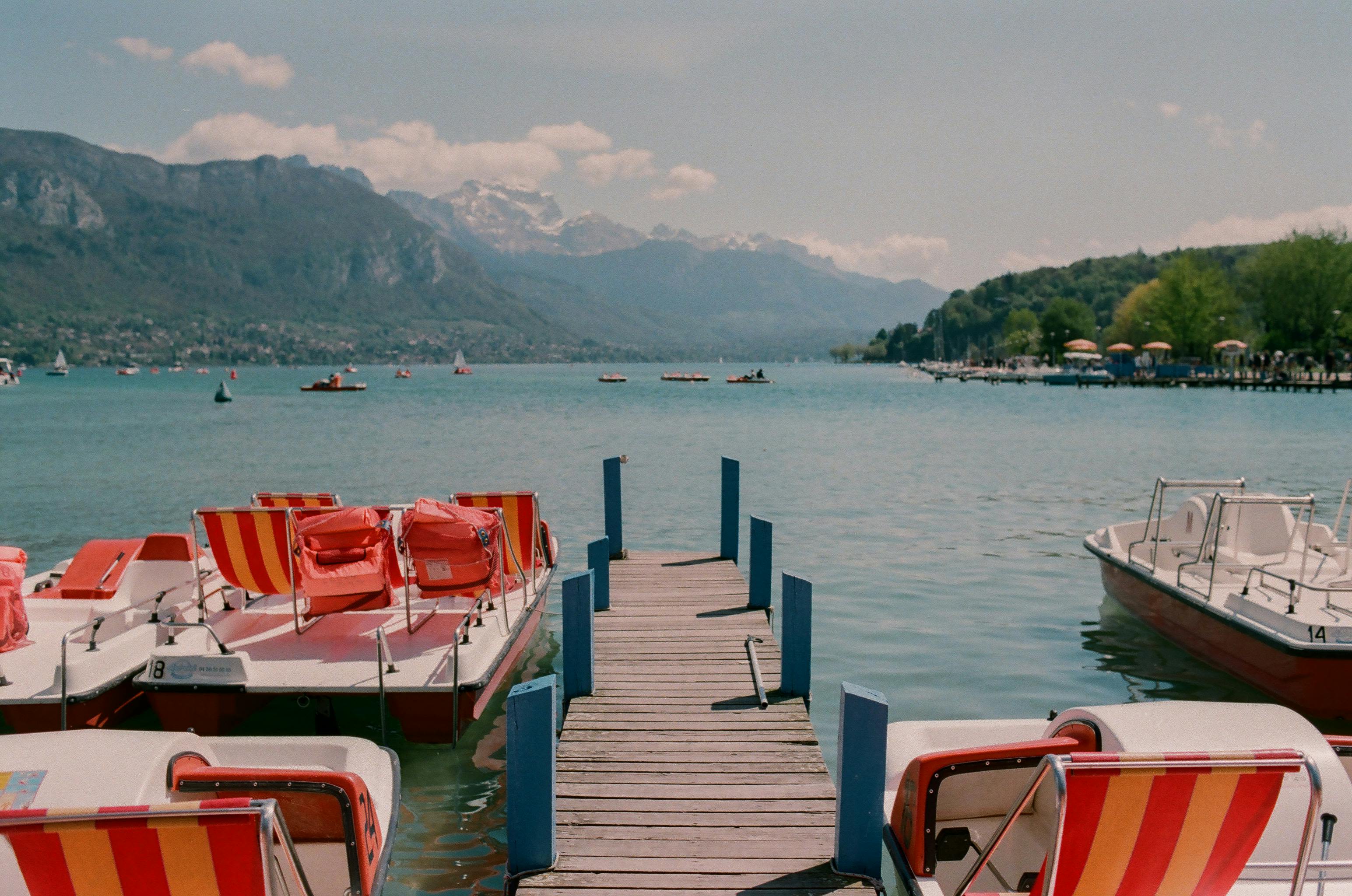 Film-style image of a boardwalk leading onto a lake with mountains in the background. Colourful boats like the path.