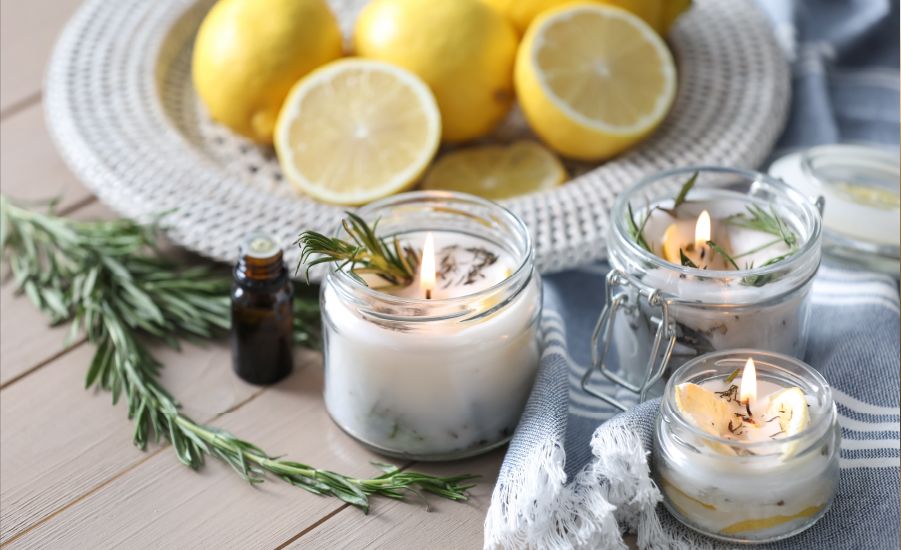 Home-made citronella candles in glass jars sit on a blue and white striped tea towel, surrounded by rosemary and essential oils. A plate of cut lemons sits in the background.