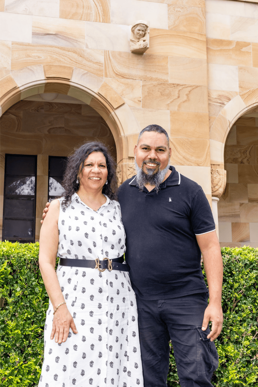 Jasmine and Scekar stand together in front of UQ's sandstone wall in the Great Court. The grotesque sits high above, looking over them.