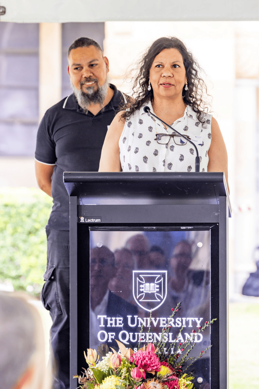 Jasmine stands before a lectern, presenting to the event attendees. Scekar, who has just spoken, stands smiling behind her.