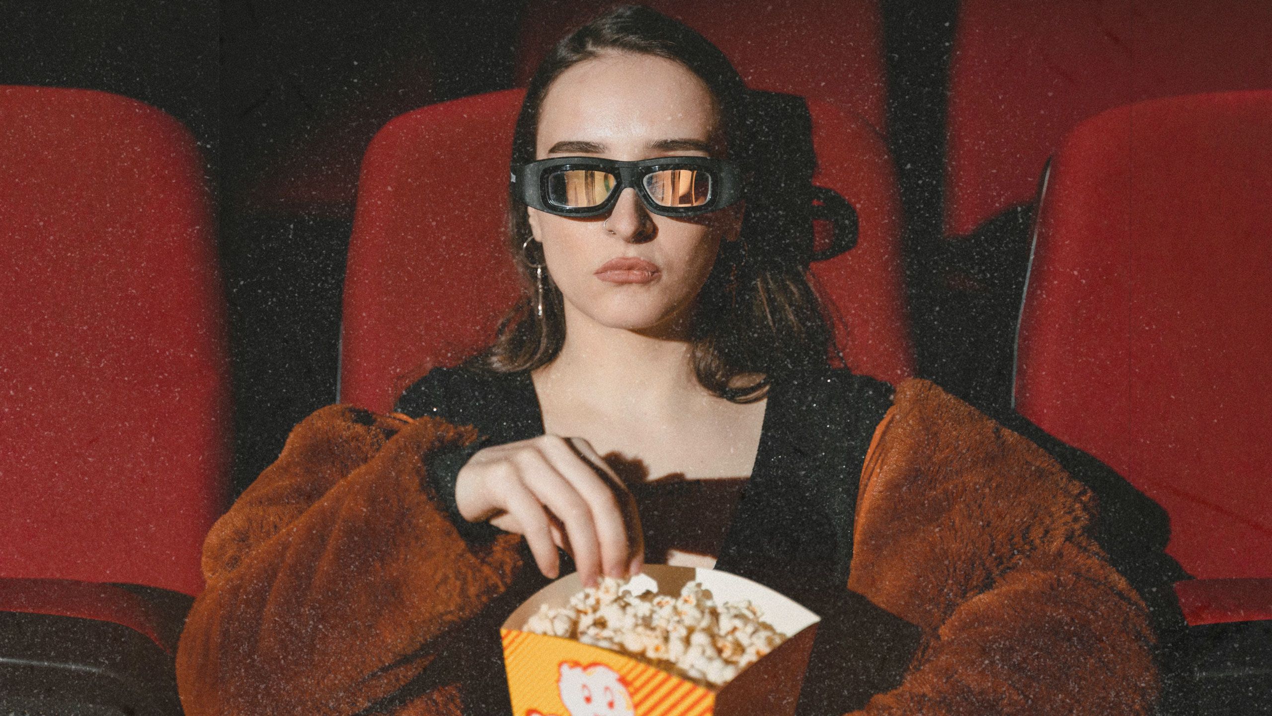 A Gen Z woman sits in a red movie cinema chair eating popcorn with reflective sunglasses on.