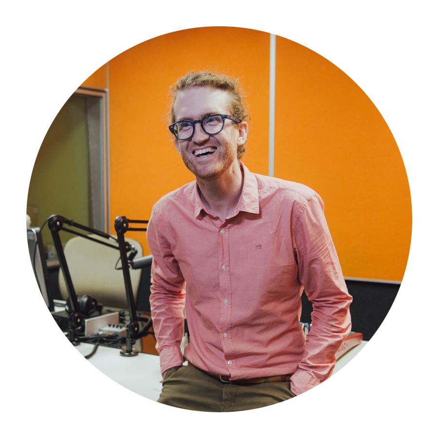 Image of an man with light skin, he wears a long sleeve pink button up shirt and dark-rimmed glasses, he is laughing and not looking at the camera