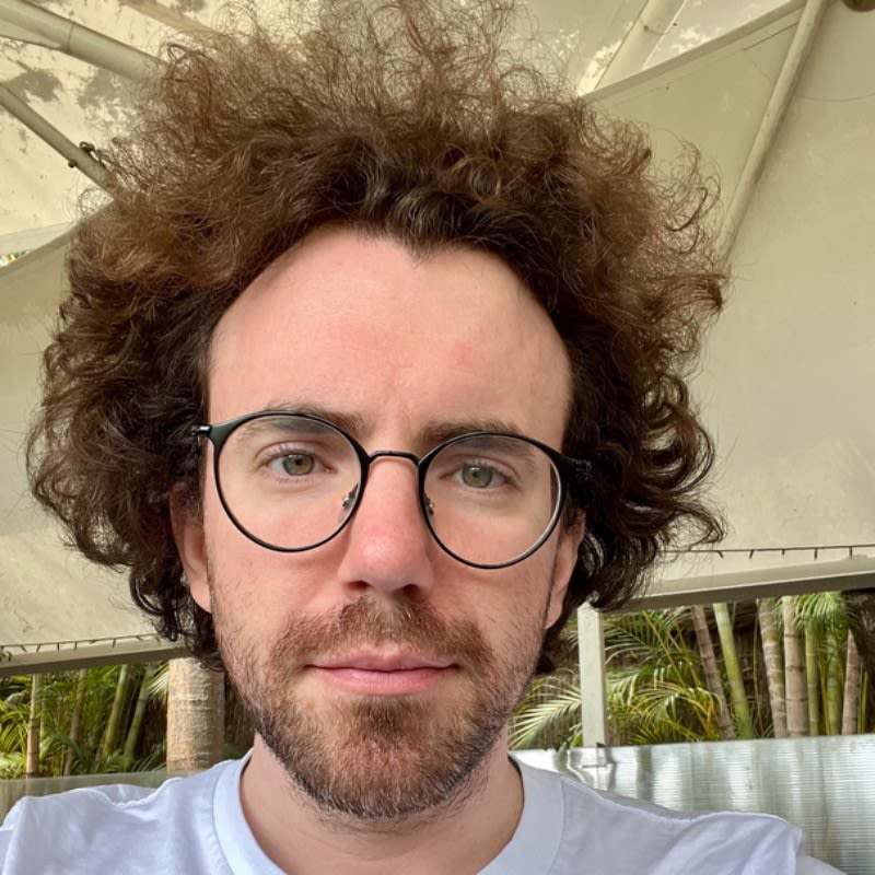 A man in his early 30s looks straight into the camera for a headshot. He has brown, very curly hair with a beard and wears round, black glasses.