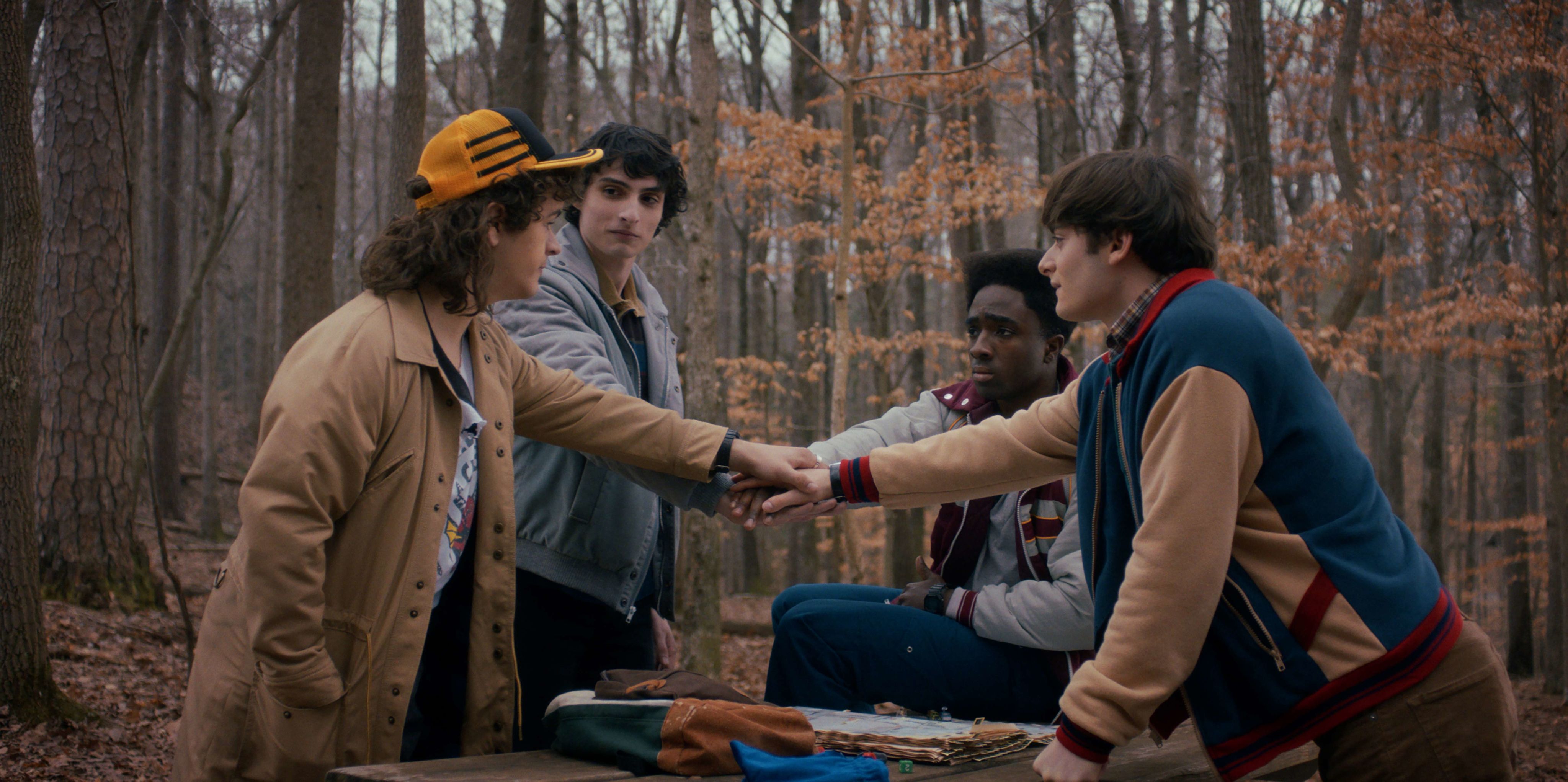 Four boys, Dustin, Mike, Lucas and Will, face each other in a forest. Their hands reach into the centre of their huddle and are placed atop each other in solidarity.
