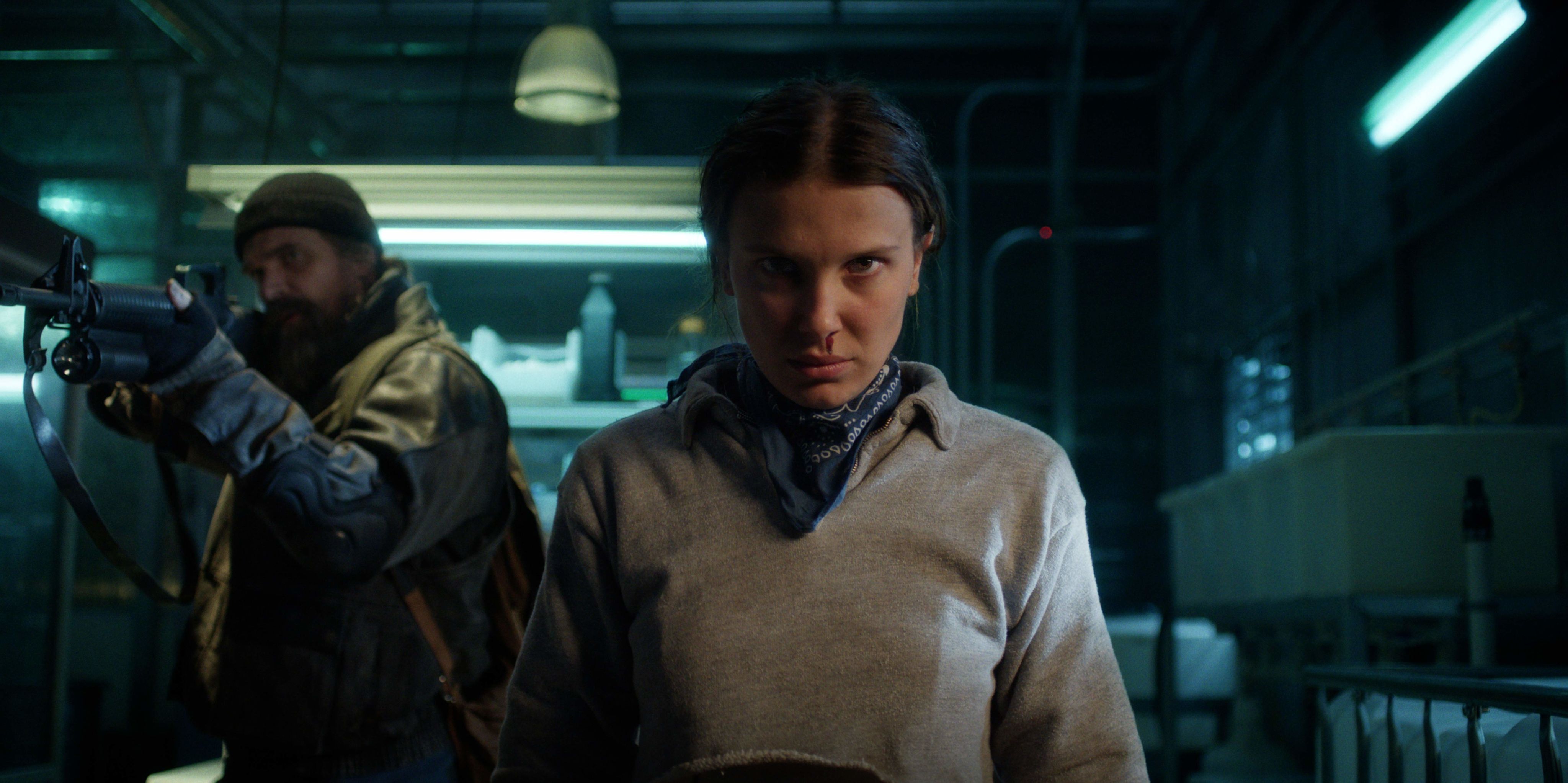 Eleven (Millie Bobbie Brown) stares straight into the camera, blood trickling out of her nose, while Jim Hopper (David Harbour) stands behind her, aiming a gun at a threat they face. 
