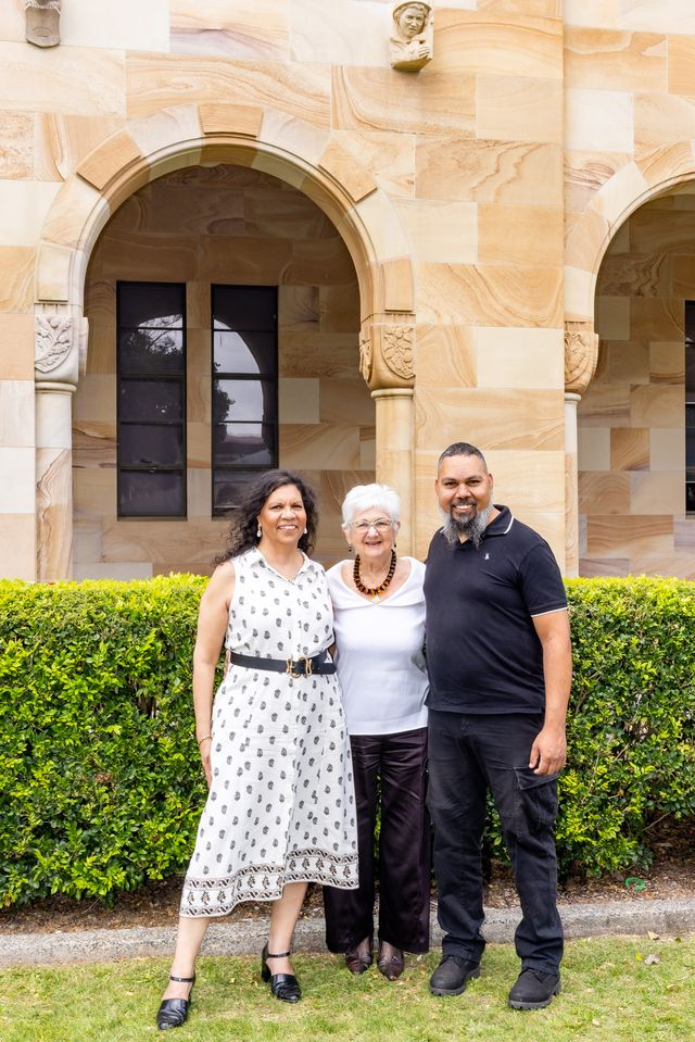 Three people standing together in front of a sandstone building with arched windows and decorative carvings, with a neatly trimmed green hedge in the foreground.