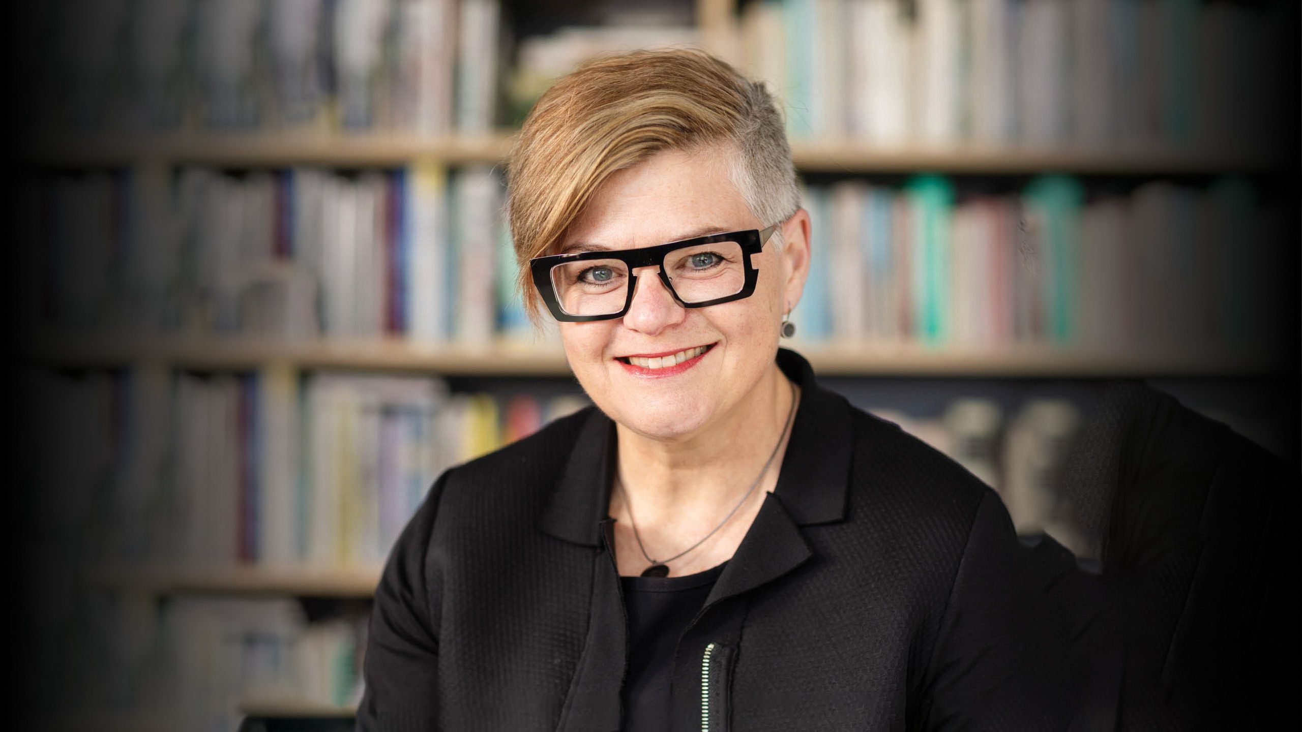 Image of a light-skinned woman in a black top, she has short cropped hair and black thick-rimmed glasses.