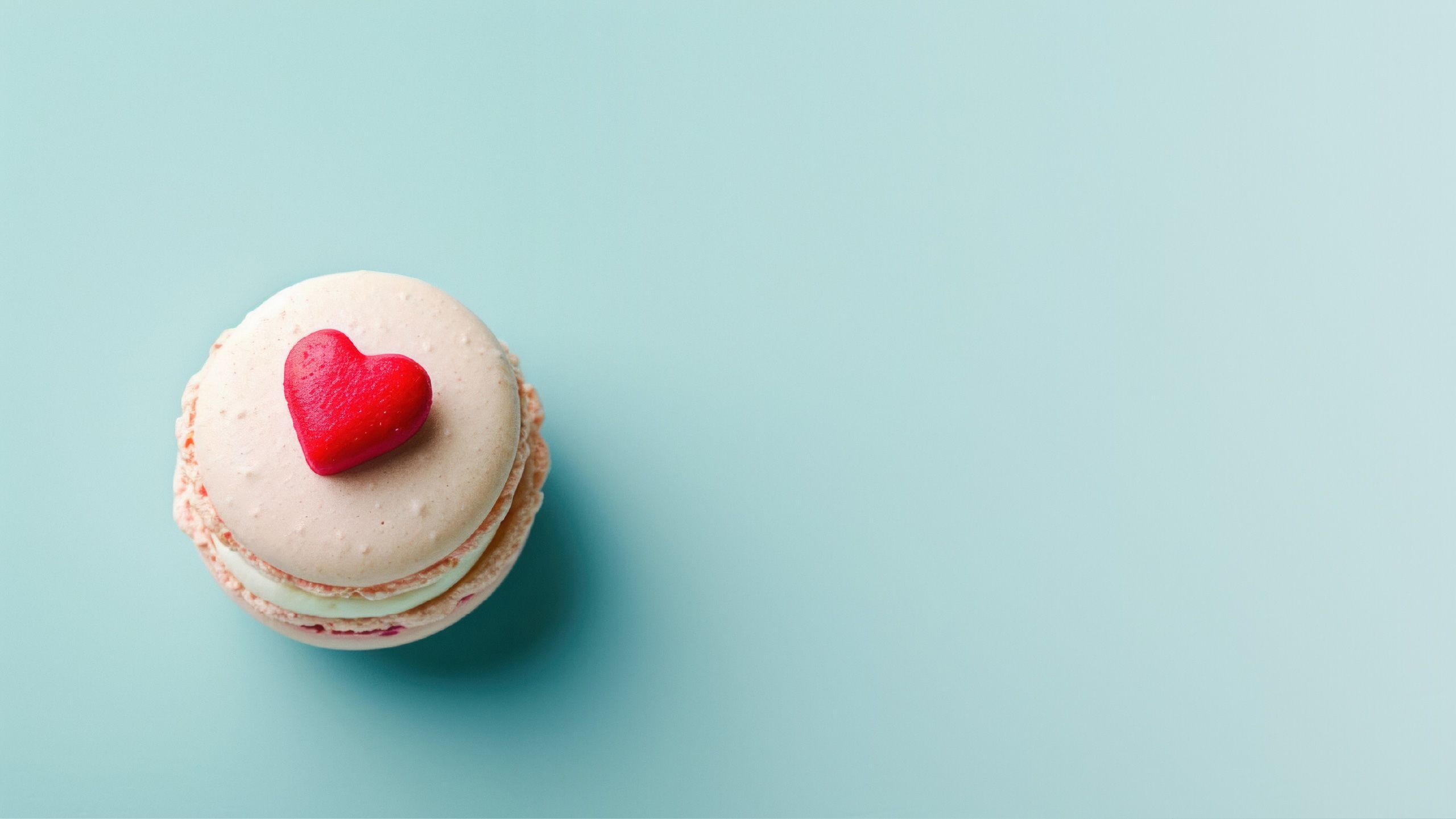 A flat lay image of a pink macaron with a red love heart on a blue background