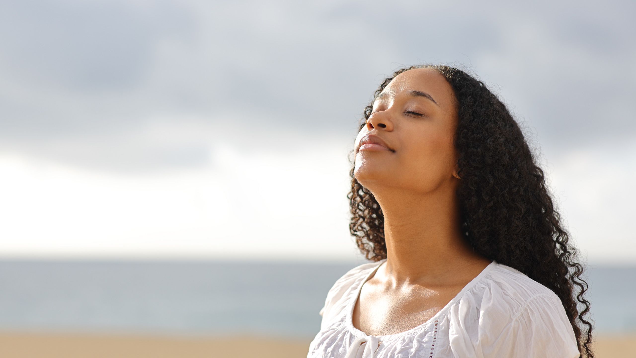 A woman stands with her head angled towards the sky and her eyes closed. The sky is cloudy but there is still some sunshine on her face.