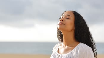 A woman stands with her head angled towards the sky and her eyes closed. The sky is cloudy but there is still some sunshine on her face.