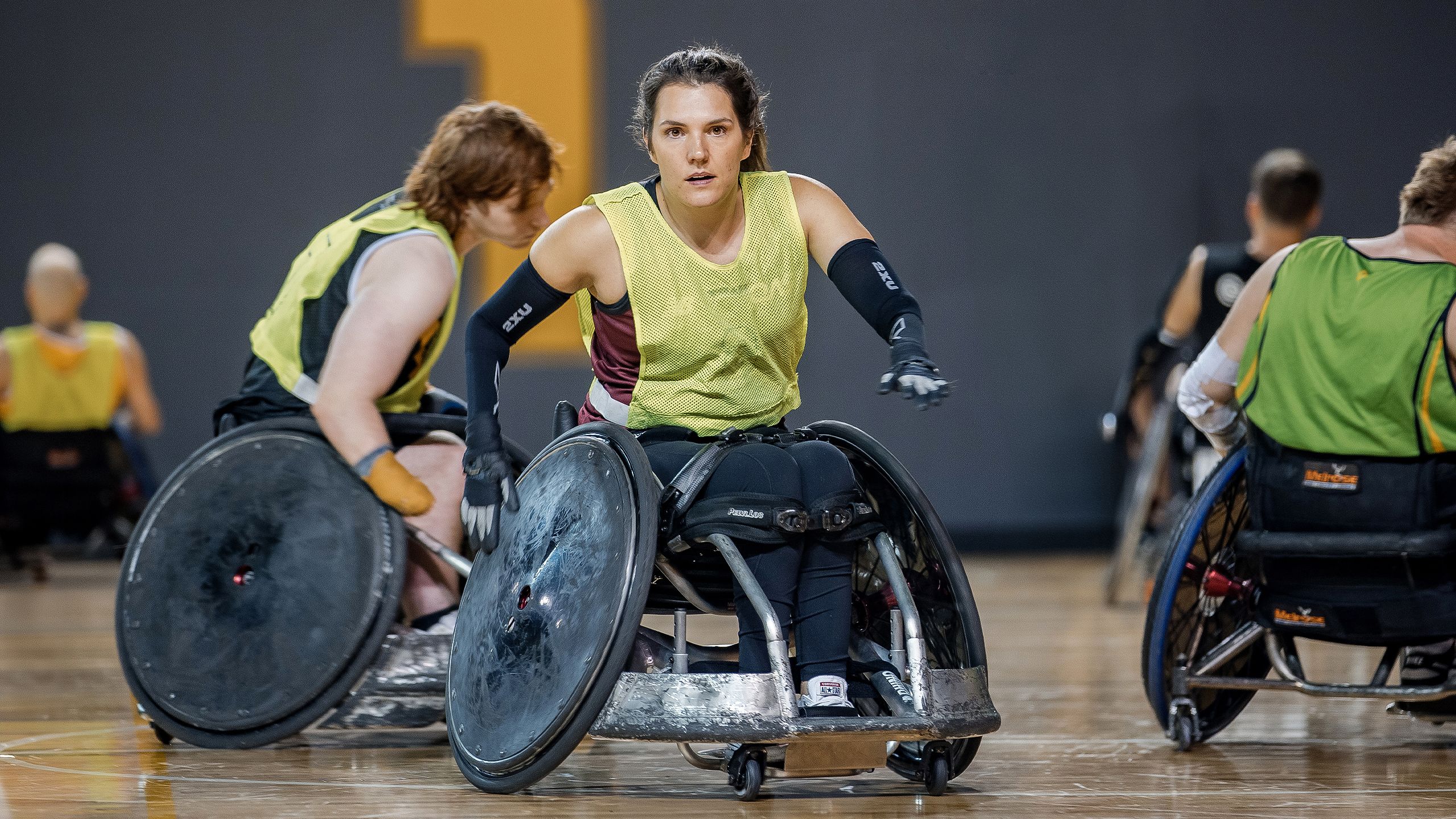 A young Para athlete in a wheelchair is on court during a wheelchair rugby game, with other players in the background.